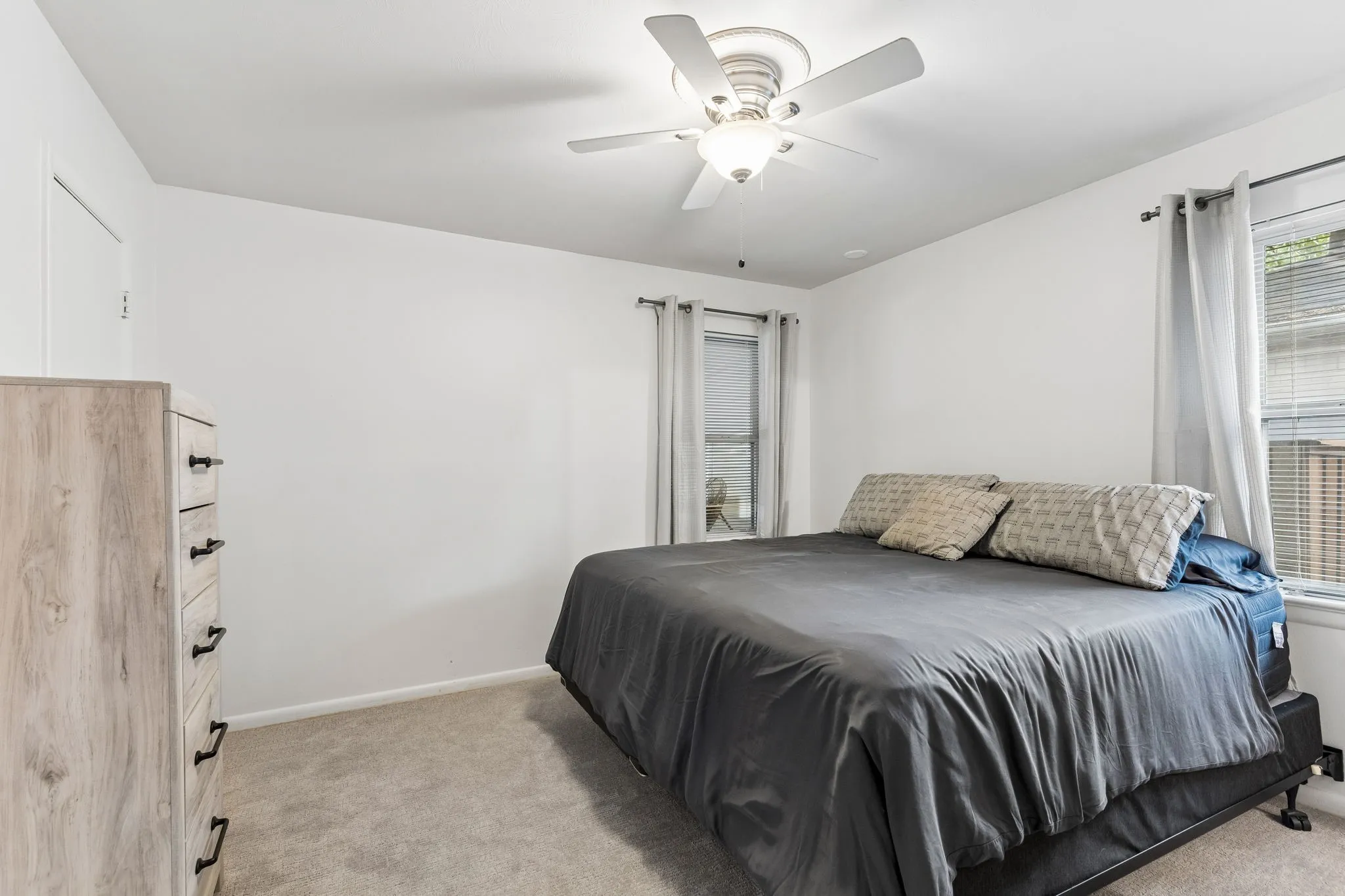 Neutral interior featuring carpeting, bright white walls, two windows with adjustable blinds, and a white ceiling-mounted fan with integrated lighting