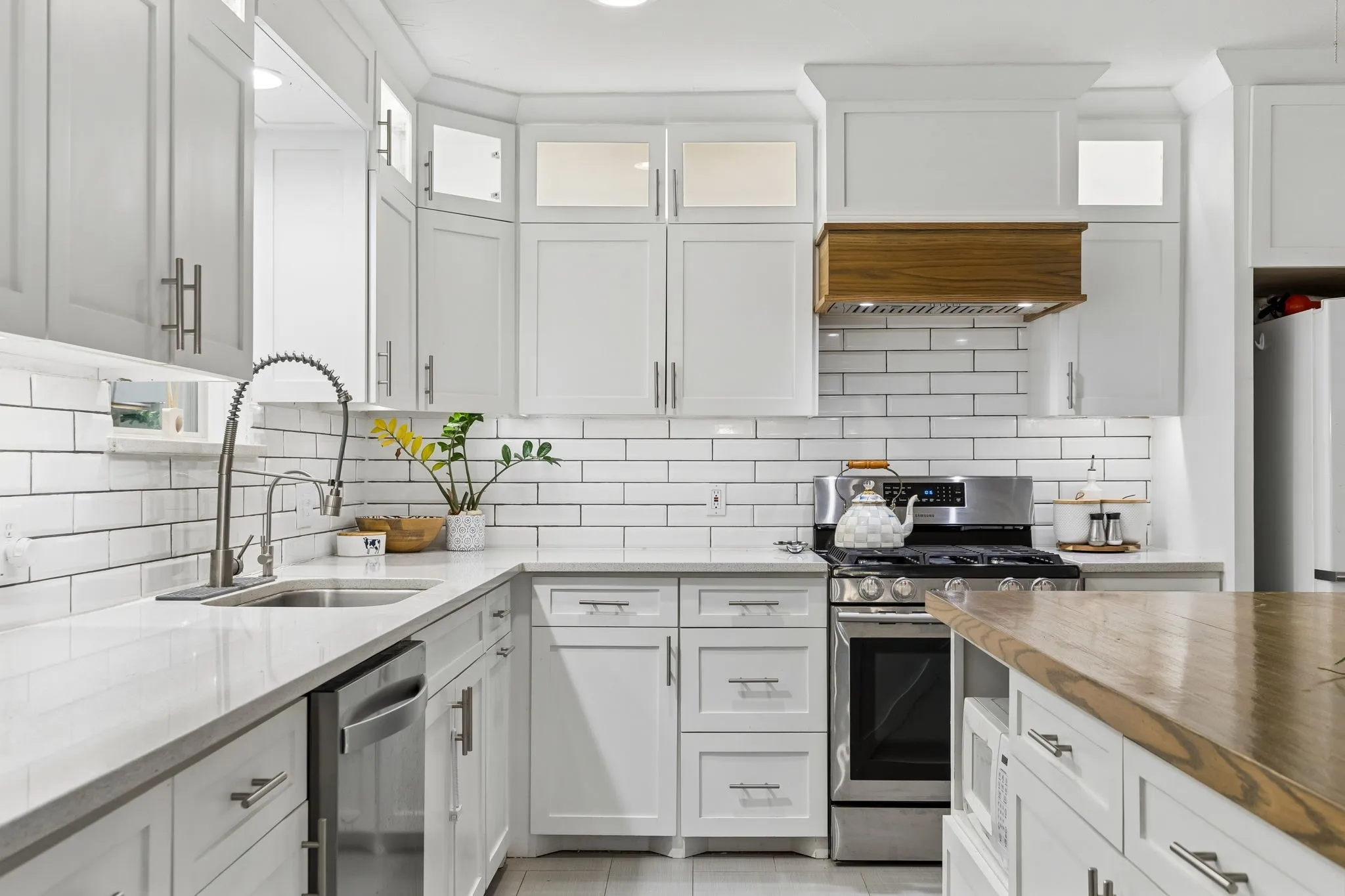 Kitchen featuring white shaker-style cabinetry with glass-front uppers, subway tile backsplash, quartz countertops, stainless steel range and dishwasher, and a custom wood-finish range hood