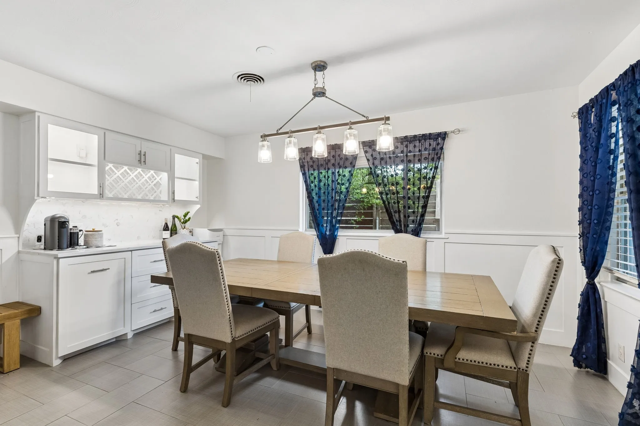 Dining area featuring white cabinetry with built-in wine storage, stone-finish countertops, ceramic tile flooring, wainscoting, and a linear pendant light fixture