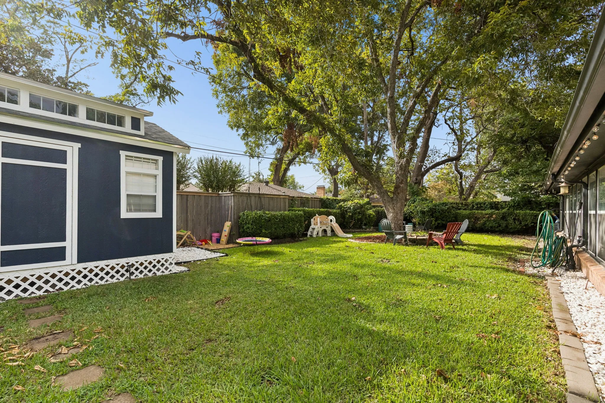 Backyard featuring a manicured lawn, perimeter wood fencing, mature trees, a detached structure with transom windows, and white gravel landscaping accents