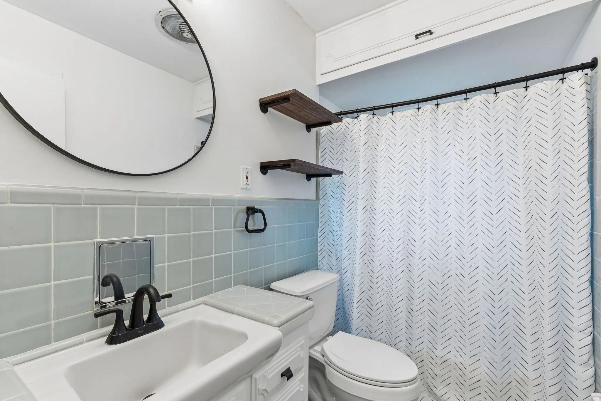 Bathroom featuring light blue square tile backsplash, white vanity with integrated sink, matte black faucet, wall-mounted circular mirror, and built-in upper cabinetry