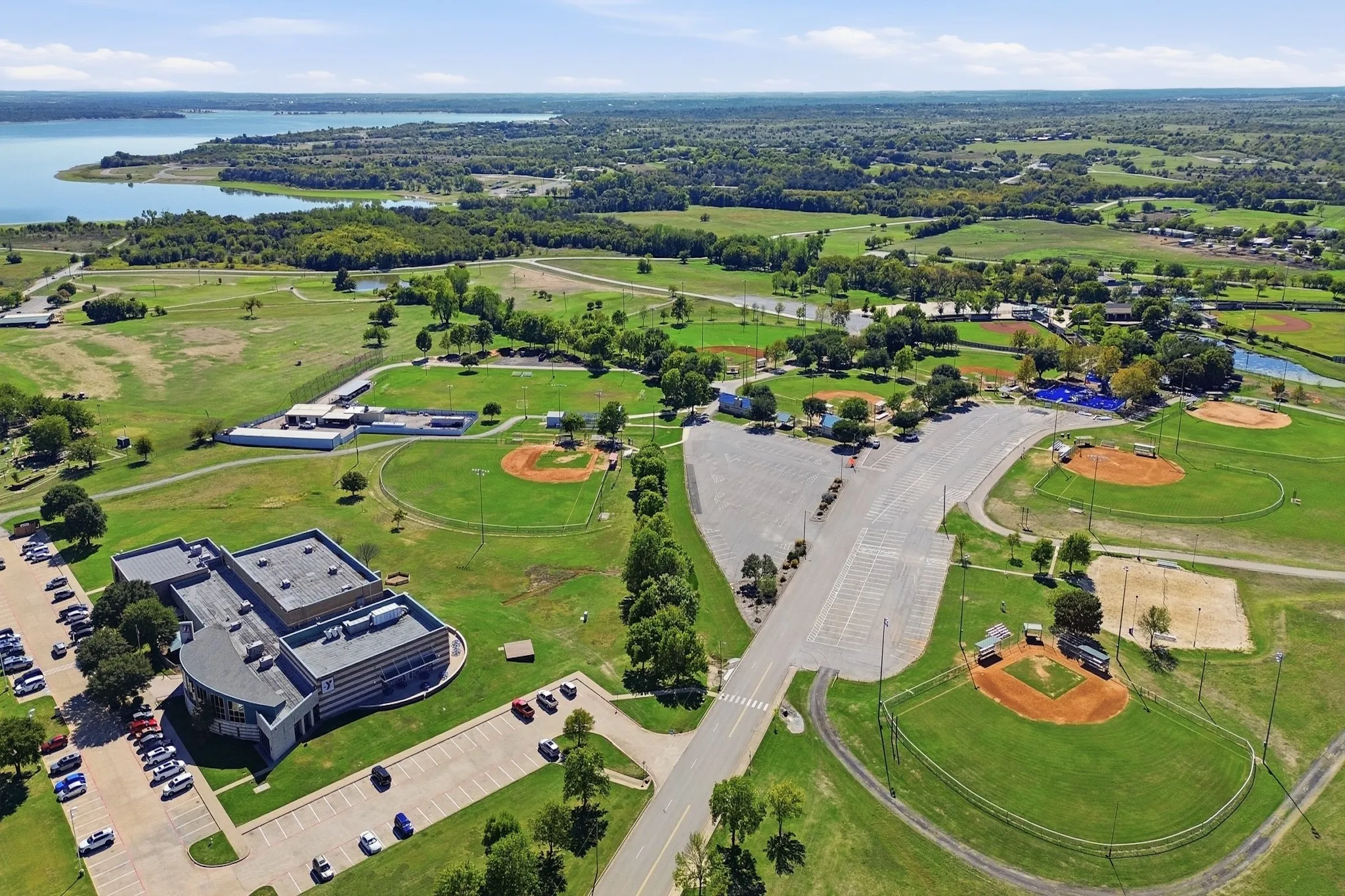 Aerial perspective of a multi-purpose recreational complex featuring multiple baseball diamonds, expansive open green space, paved parking areas, and proximity to a large body of water