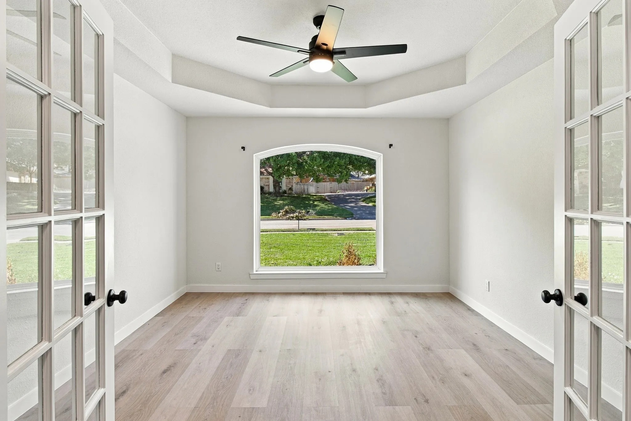 Interior space featuring French doors, wood-finish flooring, a tray ceiling, a central ceiling fan with integrated lighting, and an arched window