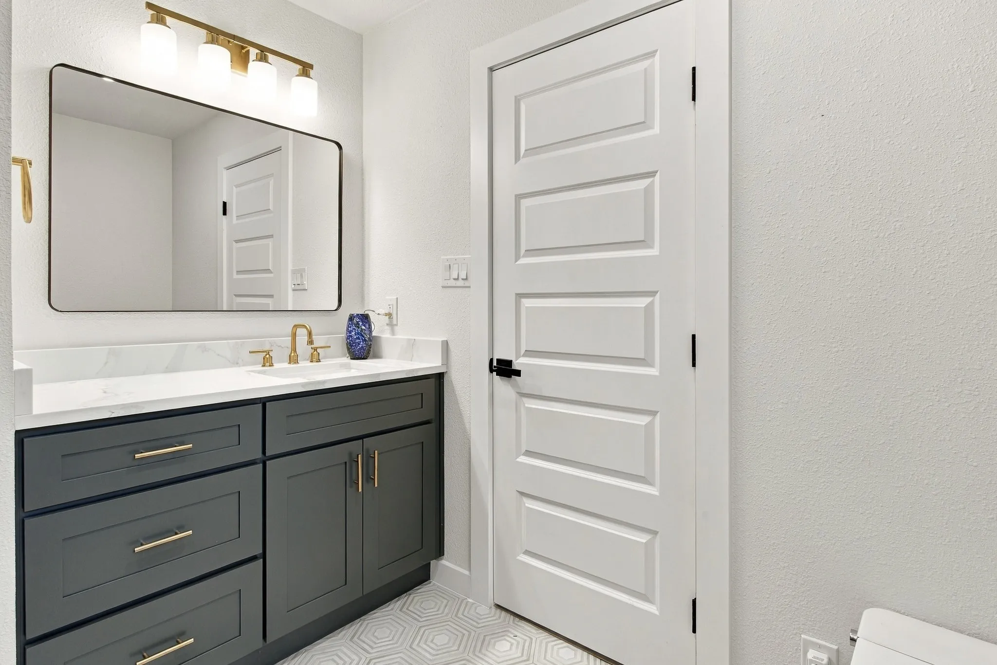 Dark grey shaker-style vanity with gold-tone hardware, white stone-finish countertop, undermount sink with gold-tone faucet, rectangular mirror with rounded corners, and multi-light vanity fixture