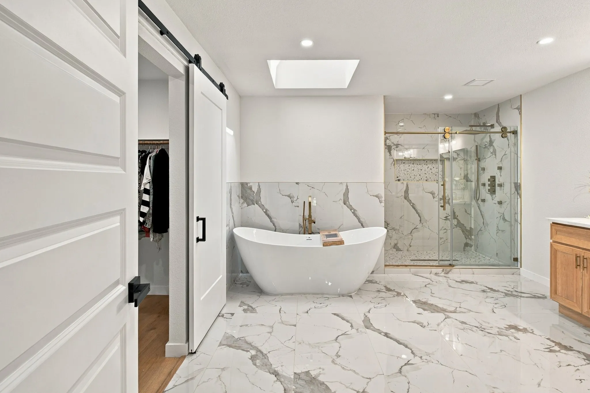 Freestanding soaking tub, walk-in shower with glass enclosure, natural stone-patterned tile flooring and wall surround, skylight, and white sliding barn door