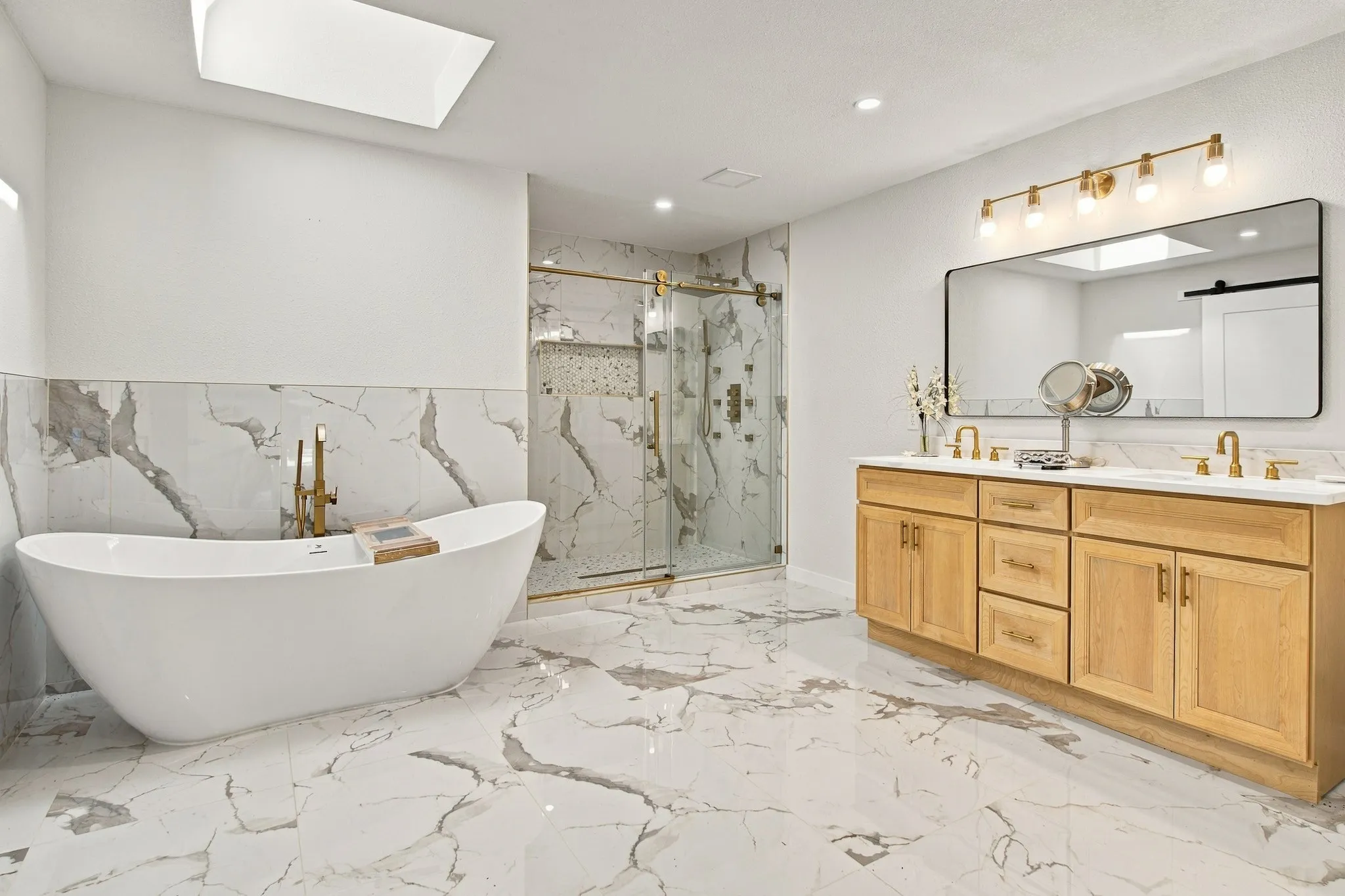 Freestanding oval soaking tub with wall-mounted gold-toned faucet, walk-in glass-enclosed shower with gold-toned fixtures, light wood cabinetry, double vanity with gold-toned hardware, and large-format porcelain tile flooring with veined detailing