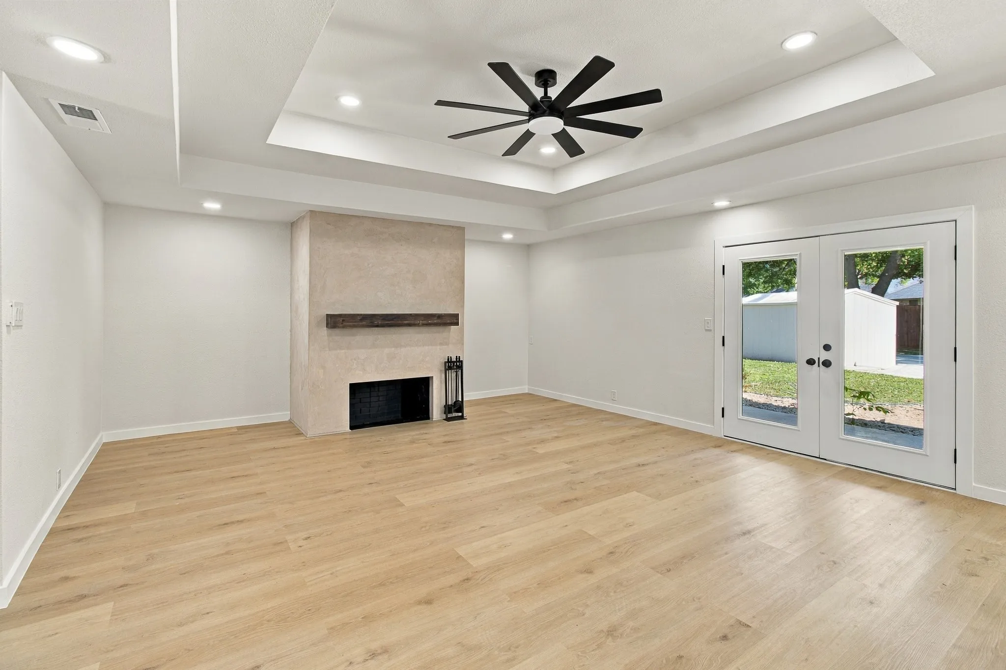 Tray ceiling featuring recessed lighting and a multi-blade ceiling fan, wood-finish flooring, masonry fireplace with a timber mantel, and glass-paneled double doors
