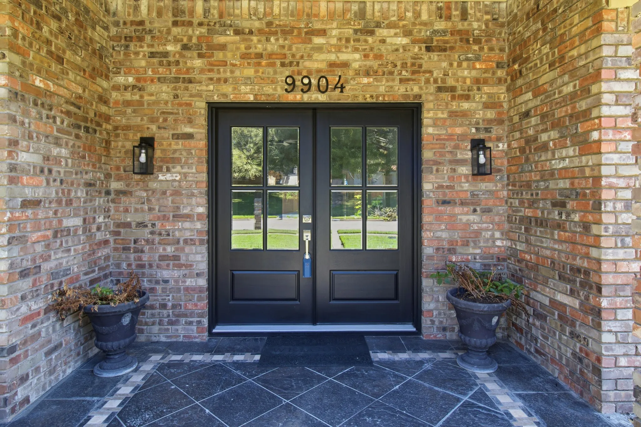 Multi-tone brick exterior featuring double front doors with glass panes, stone-tiled porch flooring with decorative border, and dual exterior wall-mounted lanterns
