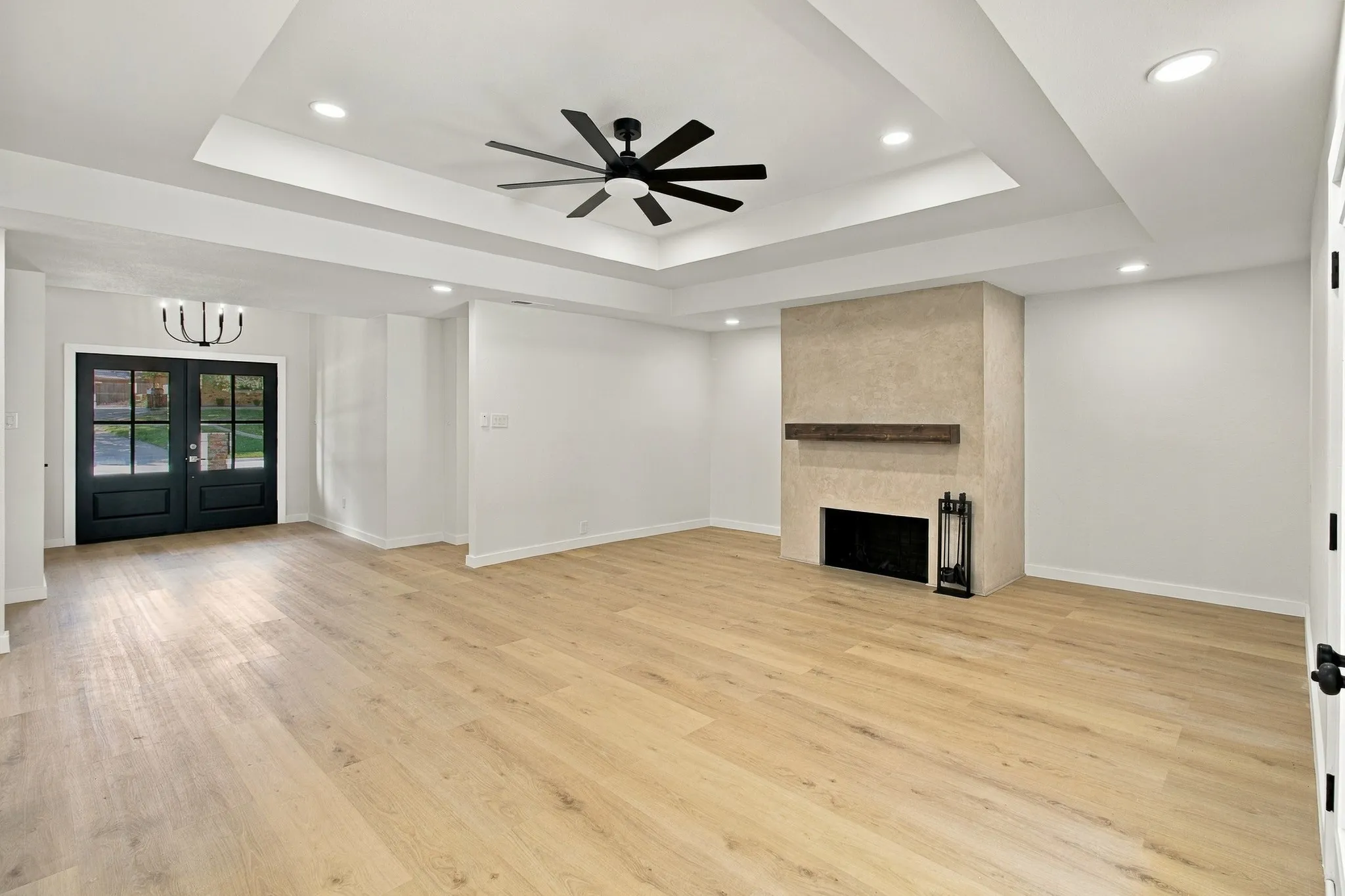 Tray ceiling featuring recessed lighting and a central ceiling fan, wood-finish flooring, and a floor-to-ceiling fireplace with a wood mantelpiece, accented by double French doors with black frames