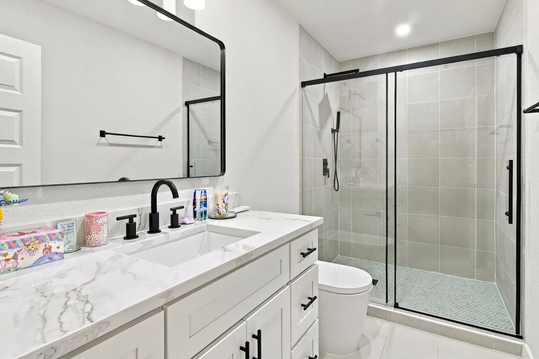 White shaker-style cabinetry with stone-finish countertop, undermount sink, matte black hardware, rectangular mirror with black frame, and a tiled walk-in shower featuring a matte black sliding glass enclosure