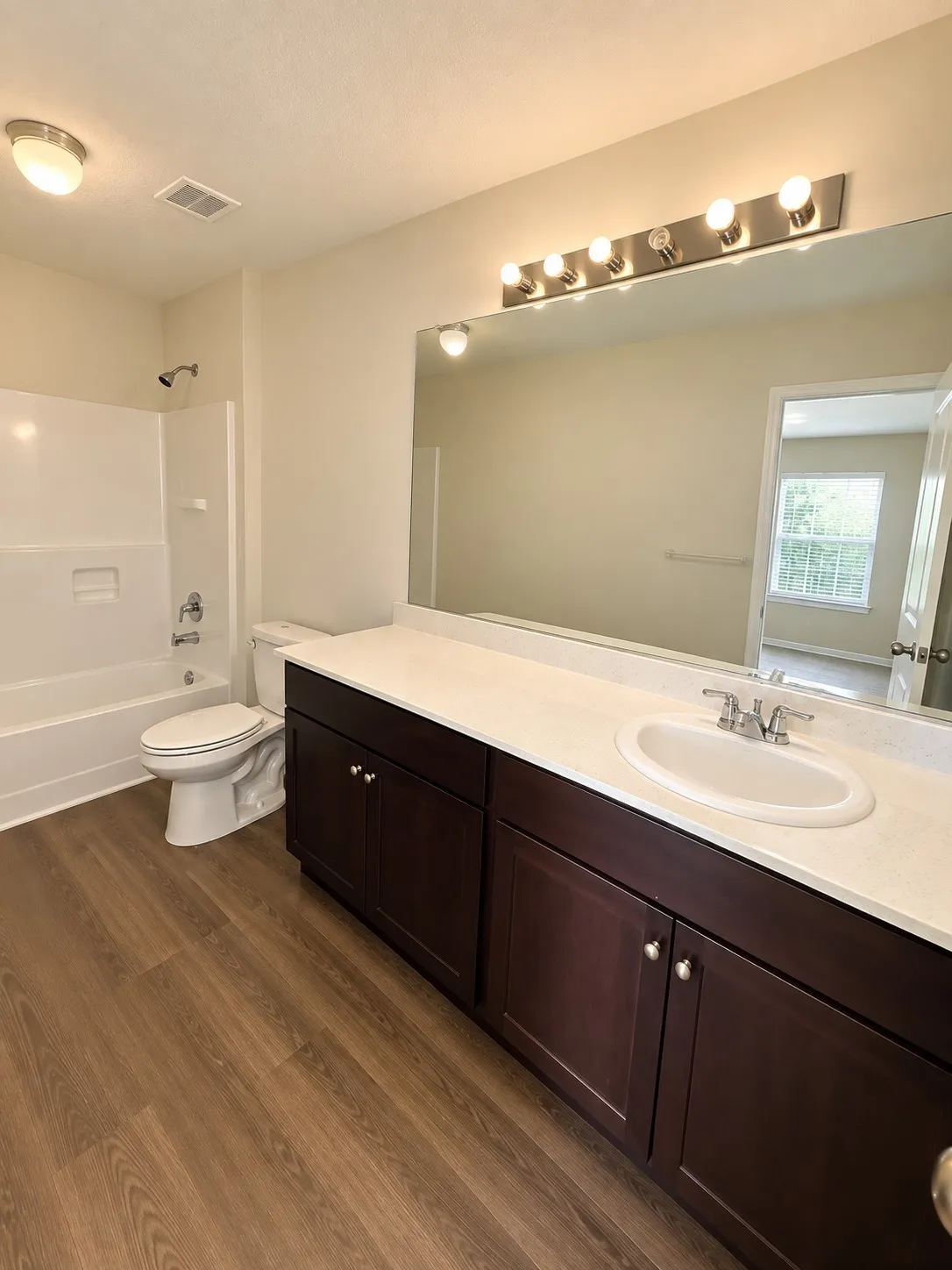 Wood-finish flooring, white vanity countertop, dark wood-finish cabinetry, single basin sink, and a shower-over-tub combination