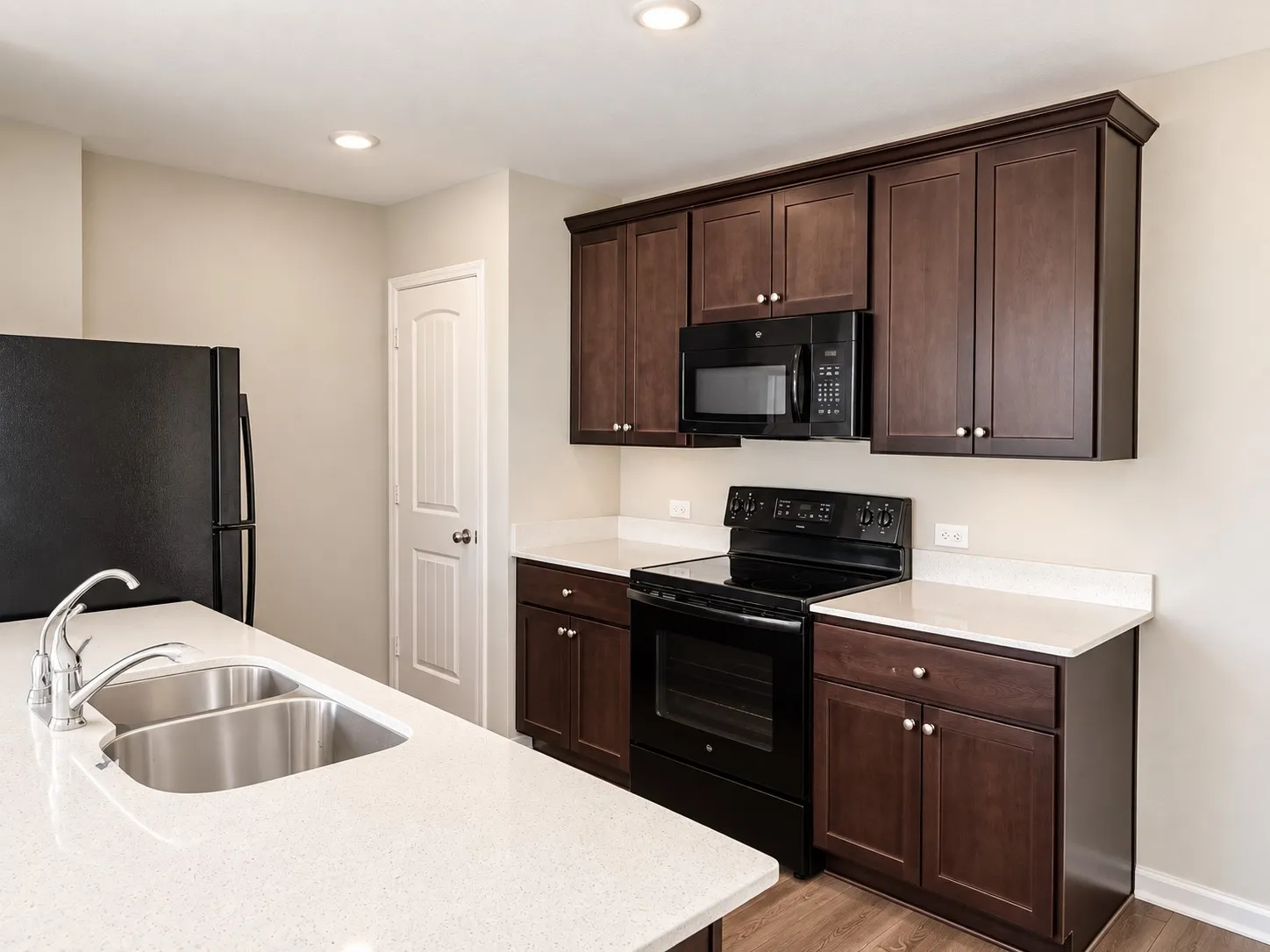 Kitchen featuring dark wood-finish cabinetry, light-toned countertops, a double basin stainless steel sink, and black appliances