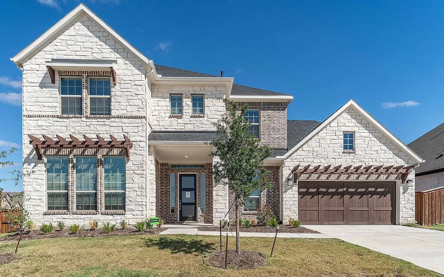 Stone and brick façade with a dark wood-finish garage door, prominent front entry, decorative window pergolas, and a gabled roofline