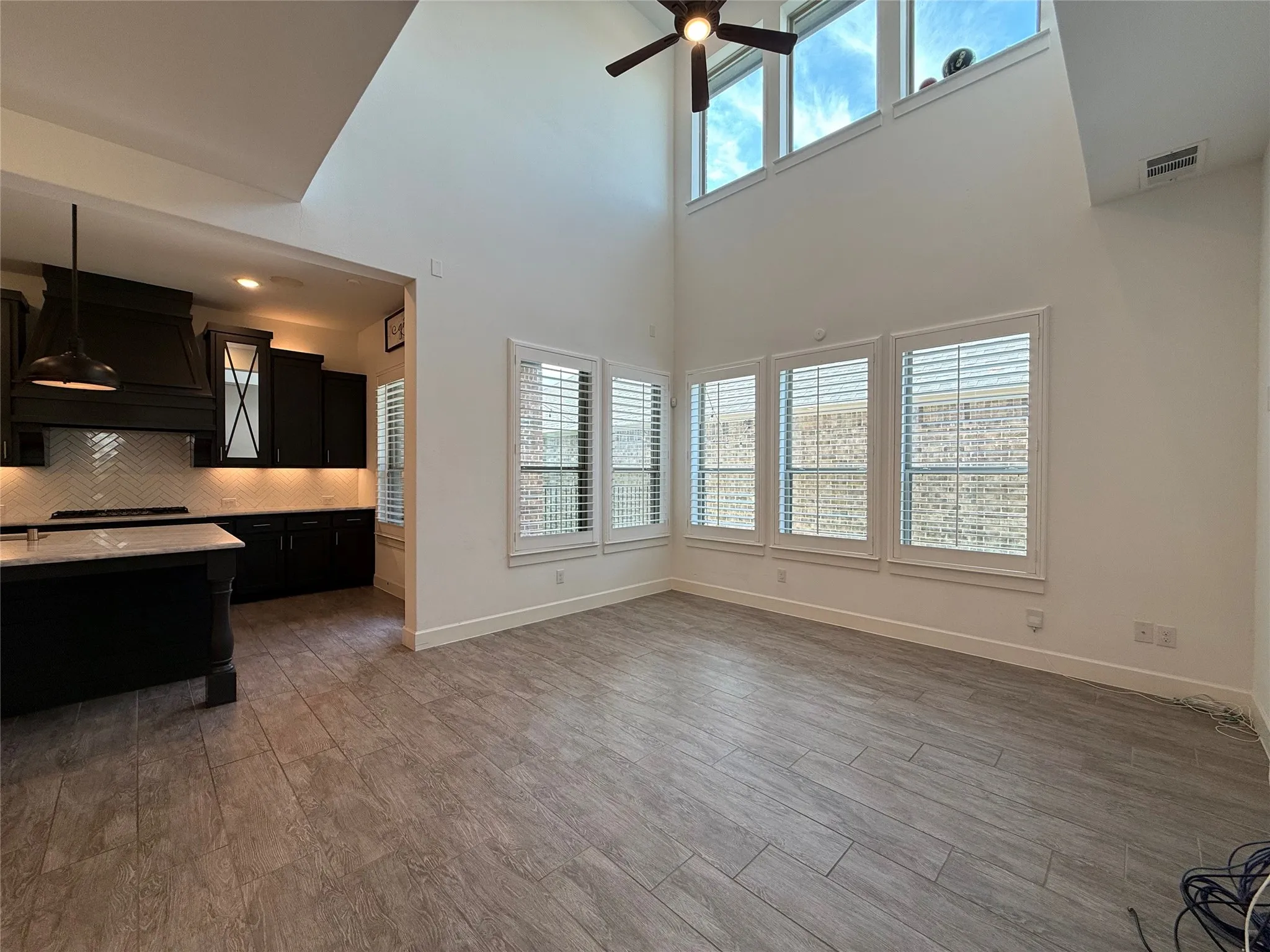 Double-height vaulted ceilings featuring clerestory windows, wood-finish tile flooring, plantation shutters, and a kitchen equipped with dark cabinetry, a center island, and a herringbone-patterned backsplash