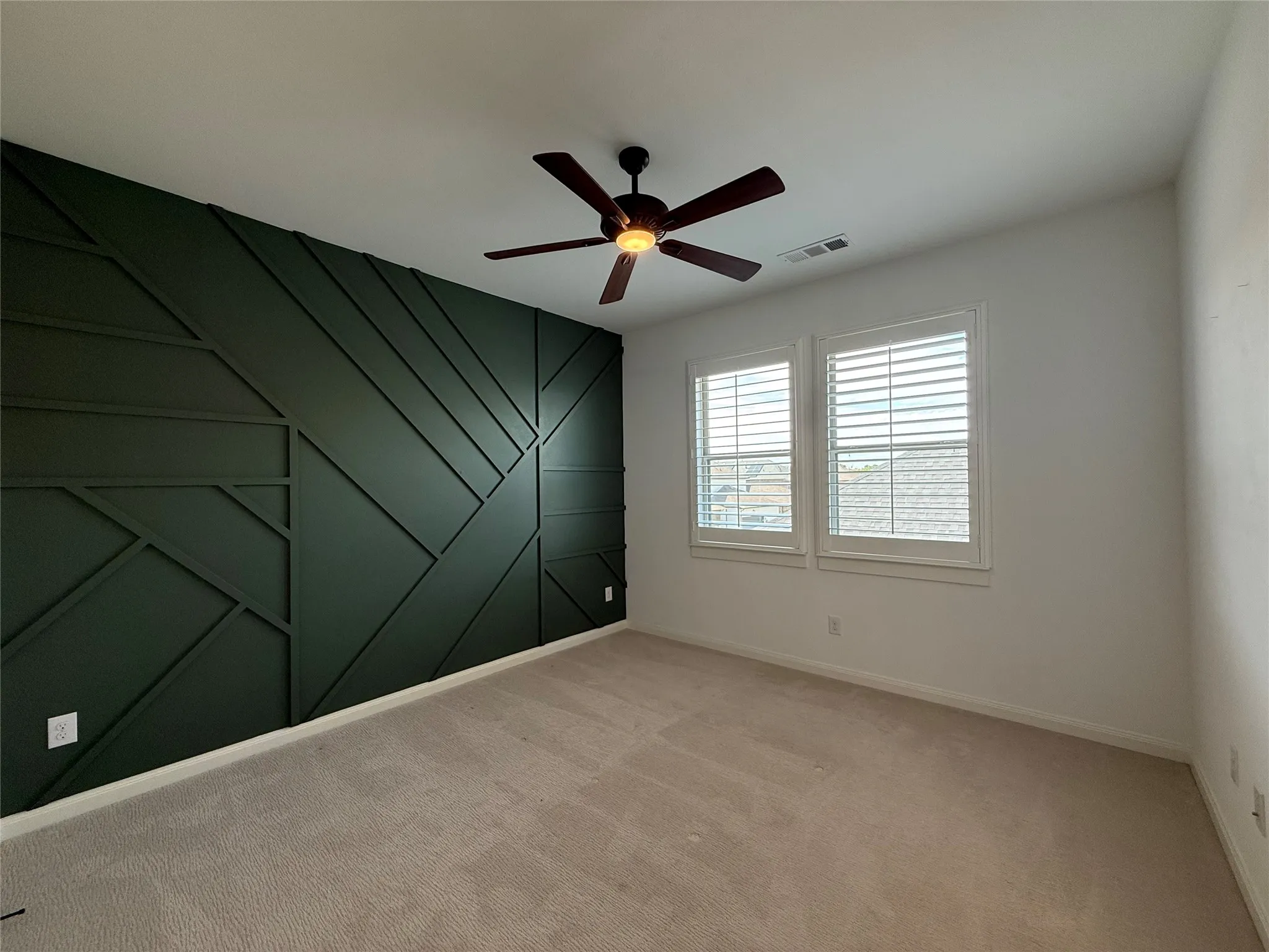 Geometric accent wall in dark finish, neutral carpeting, and shutter-style window coverings, accented by a ceiling fan with integrated light fixture