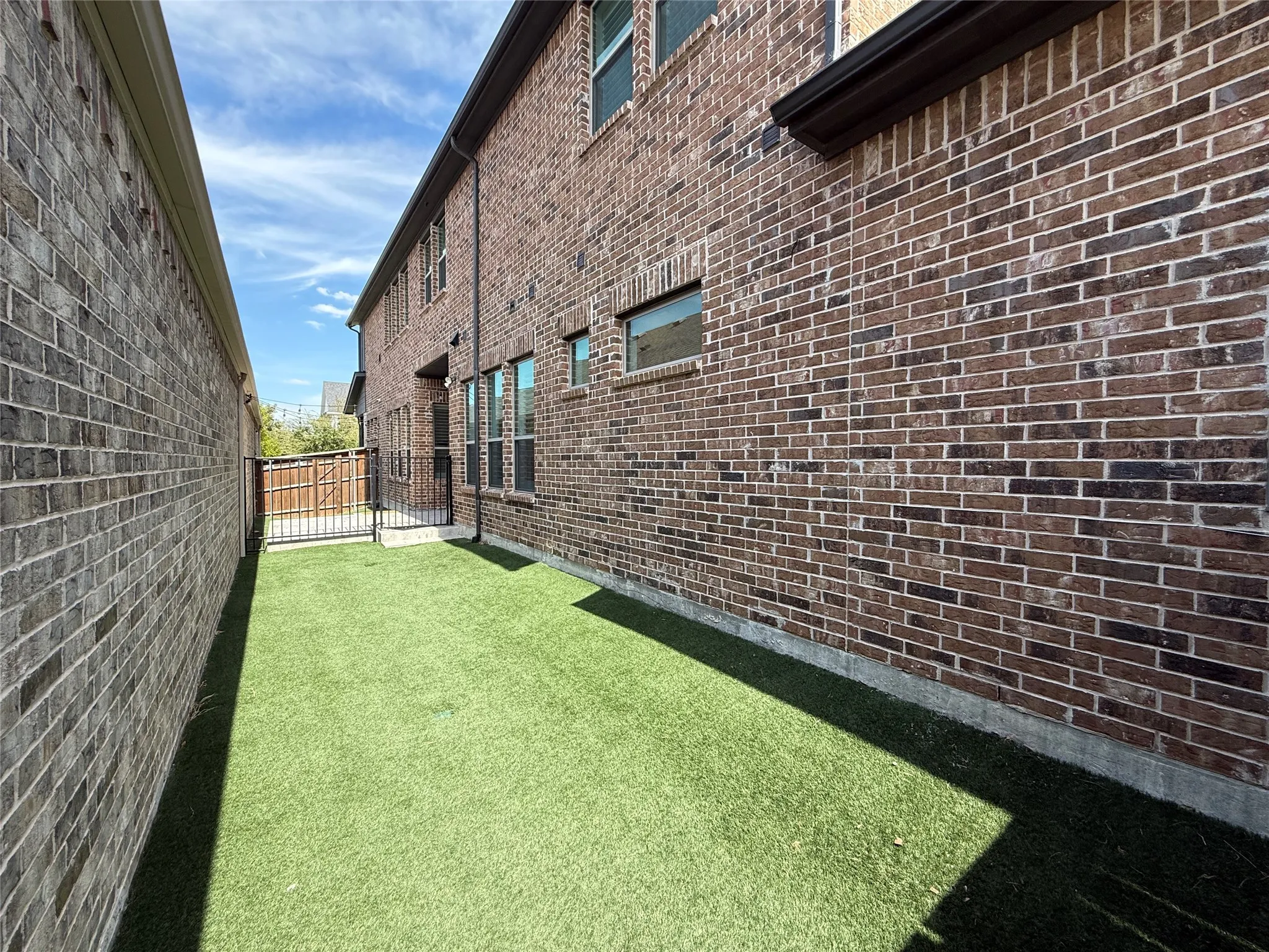 Exterior side yard featuring brick masonry construction, synthetic turf, and a metal security gate