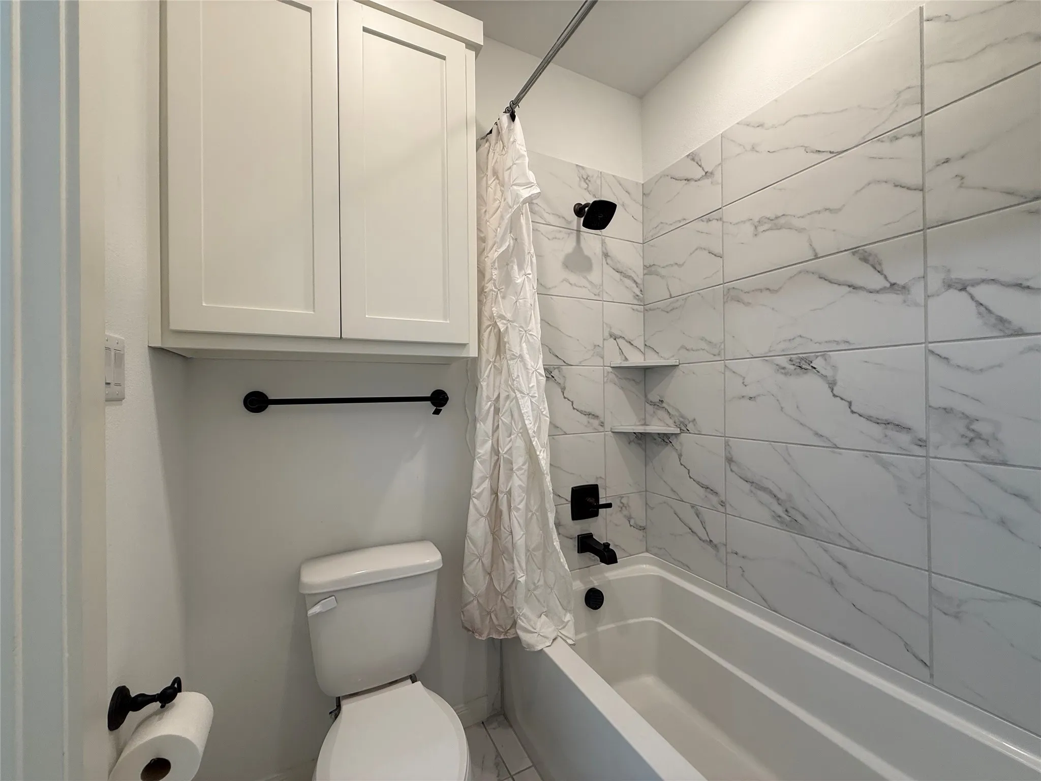 Full bathroom featuring white shaker-style wall cabinetry, large-format tile surround with integrated shelving, and matte black plumbing fixtures