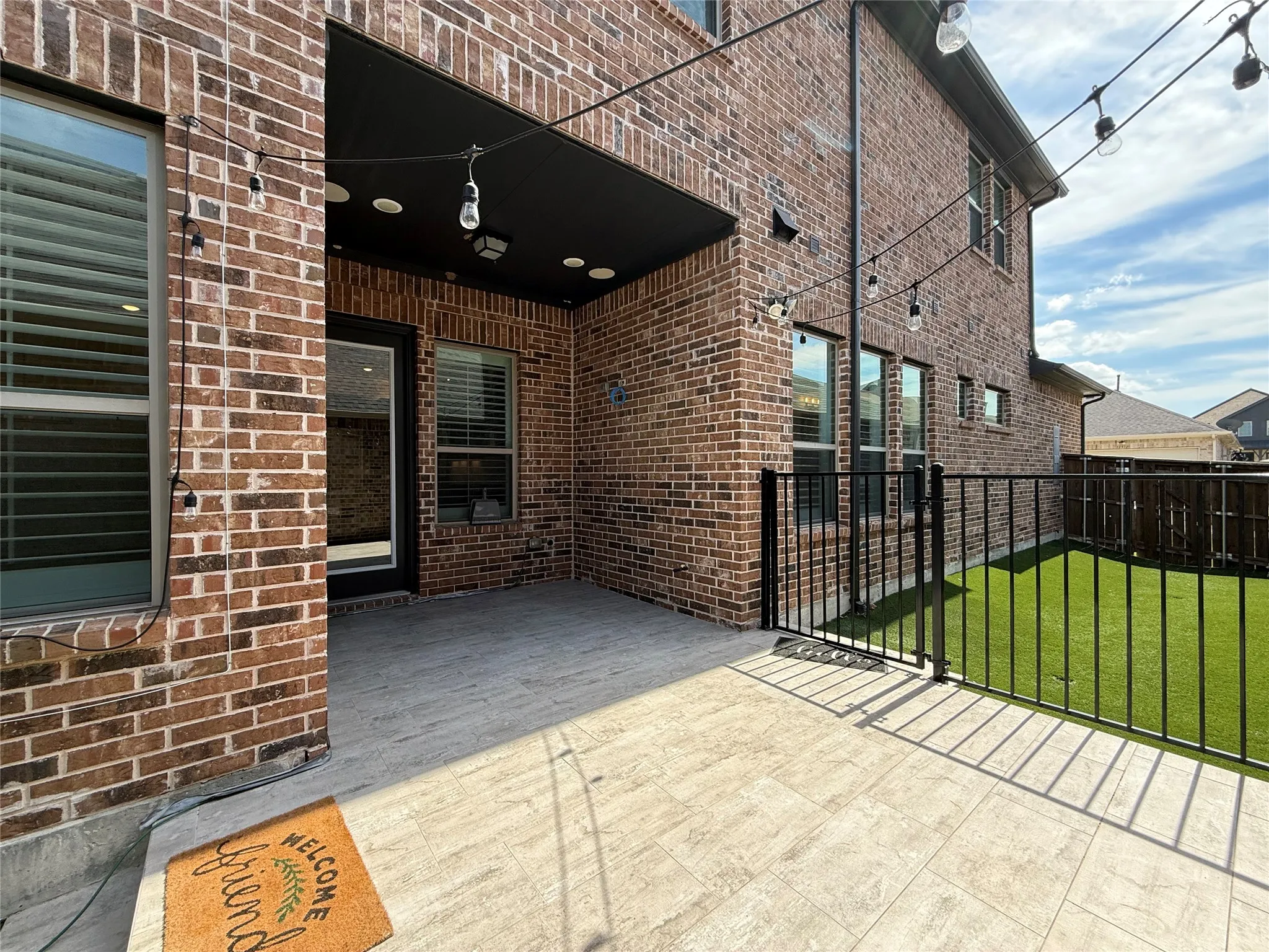 Covered brick patio featuring stone-tile flooring, recessed lighting, and a black metal perimeter fence enclosing an artificial turf lawn