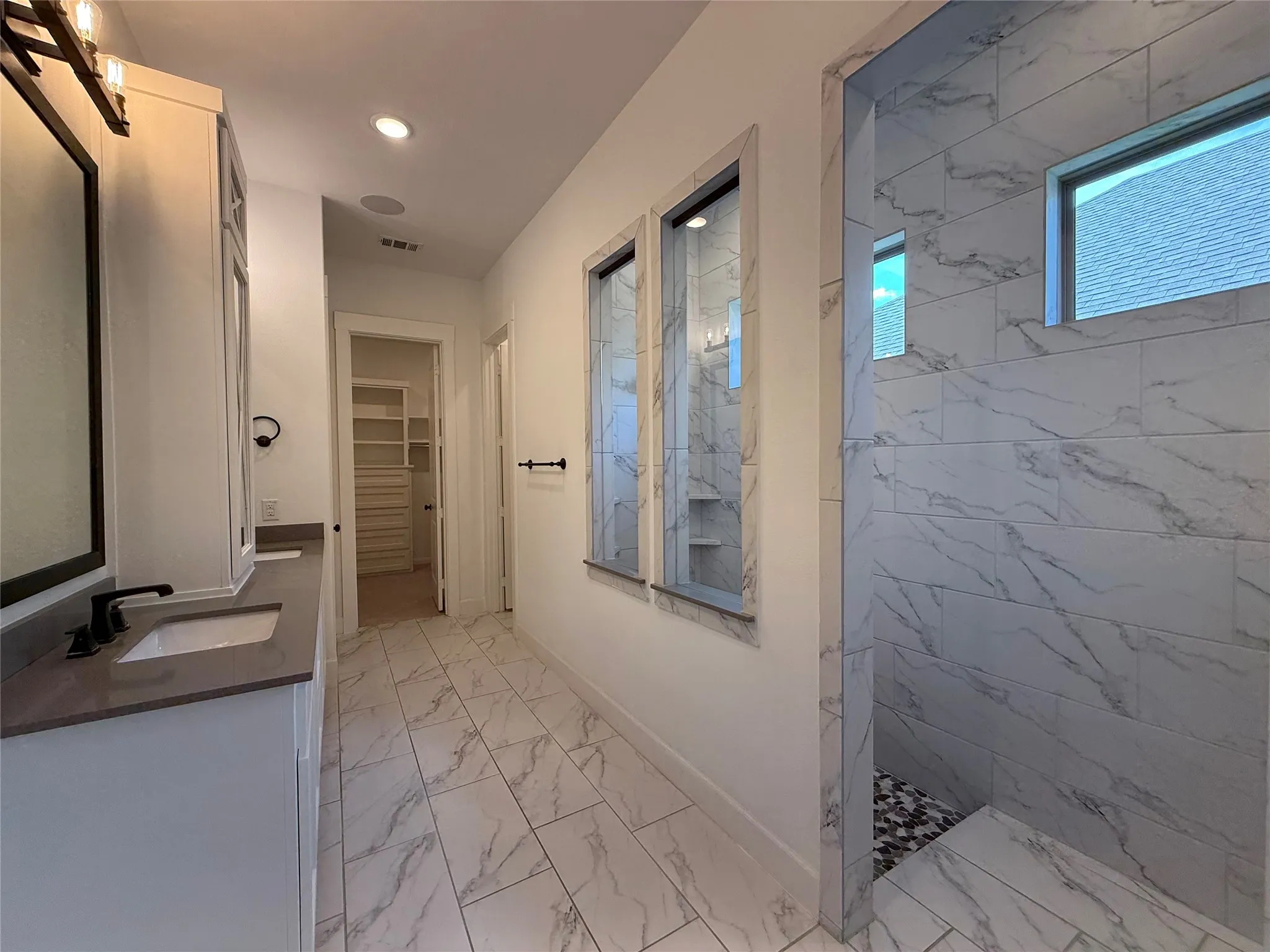Dual vanity featuring solid-surface countertops, walk-in shower with stone-patterned tile, porcelain tile flooring, and integrated glass partitioning
