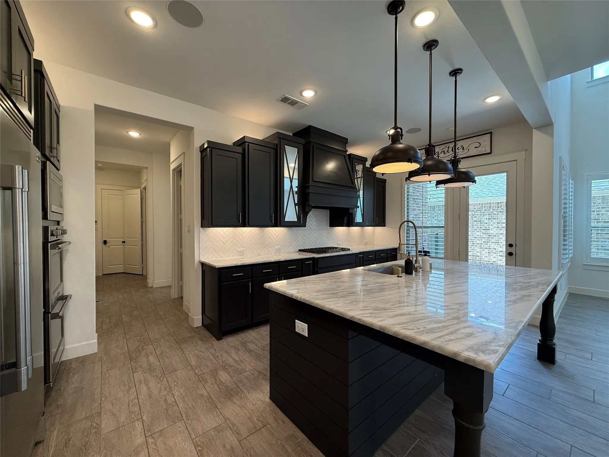 Kitchen featuring dark cabinetry, expansive stone-topped island with seating, herringbone tile backsplash, stainless steel built-in double ovens, and suspended industrial-style pendant lighting