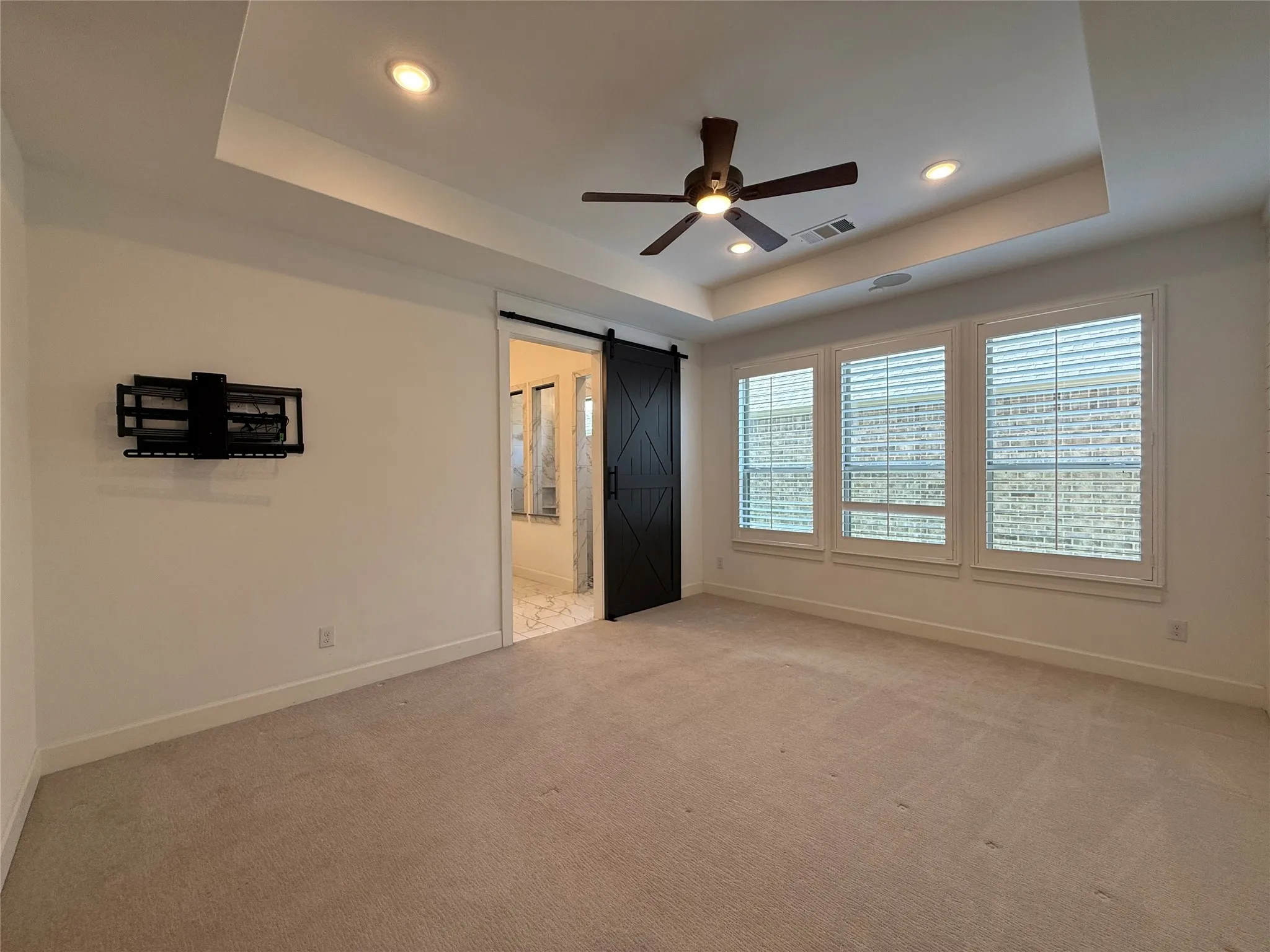 Tray ceiling with recessed lighting, black barn door, neutral carpeting, and plantation shutters