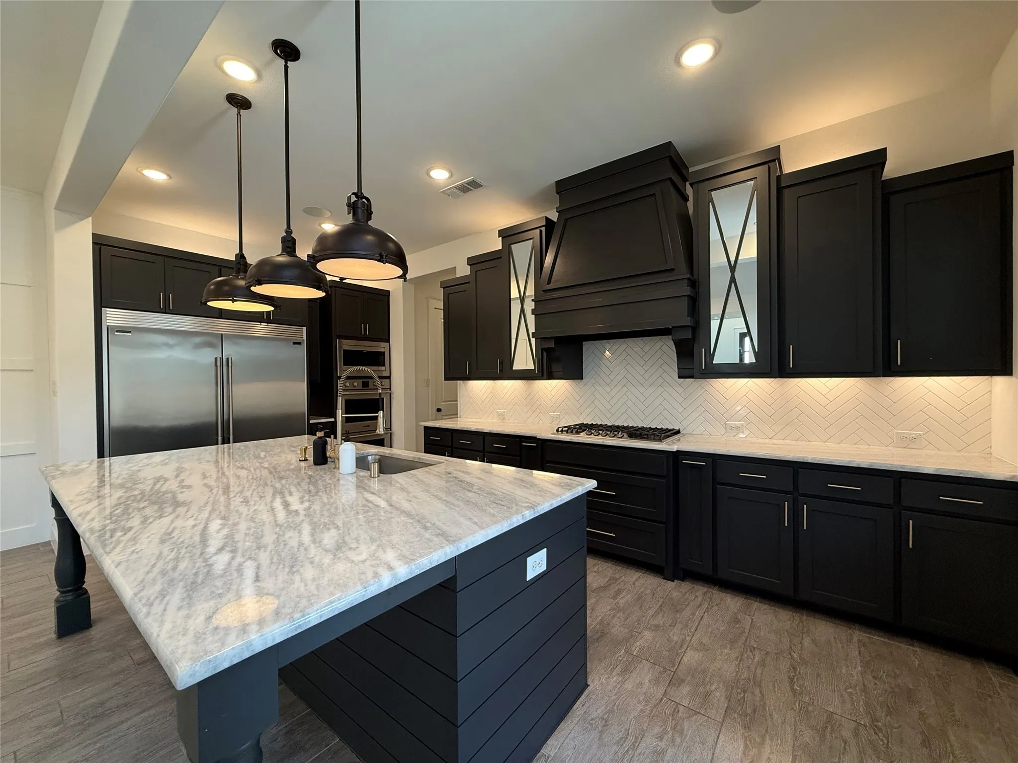 Kitchen featuring dark cabinetry, expansive stone-topped island with integrated seating, stainless steel appliances, herringbone tile backsplash, and oversized metal pendant lighting