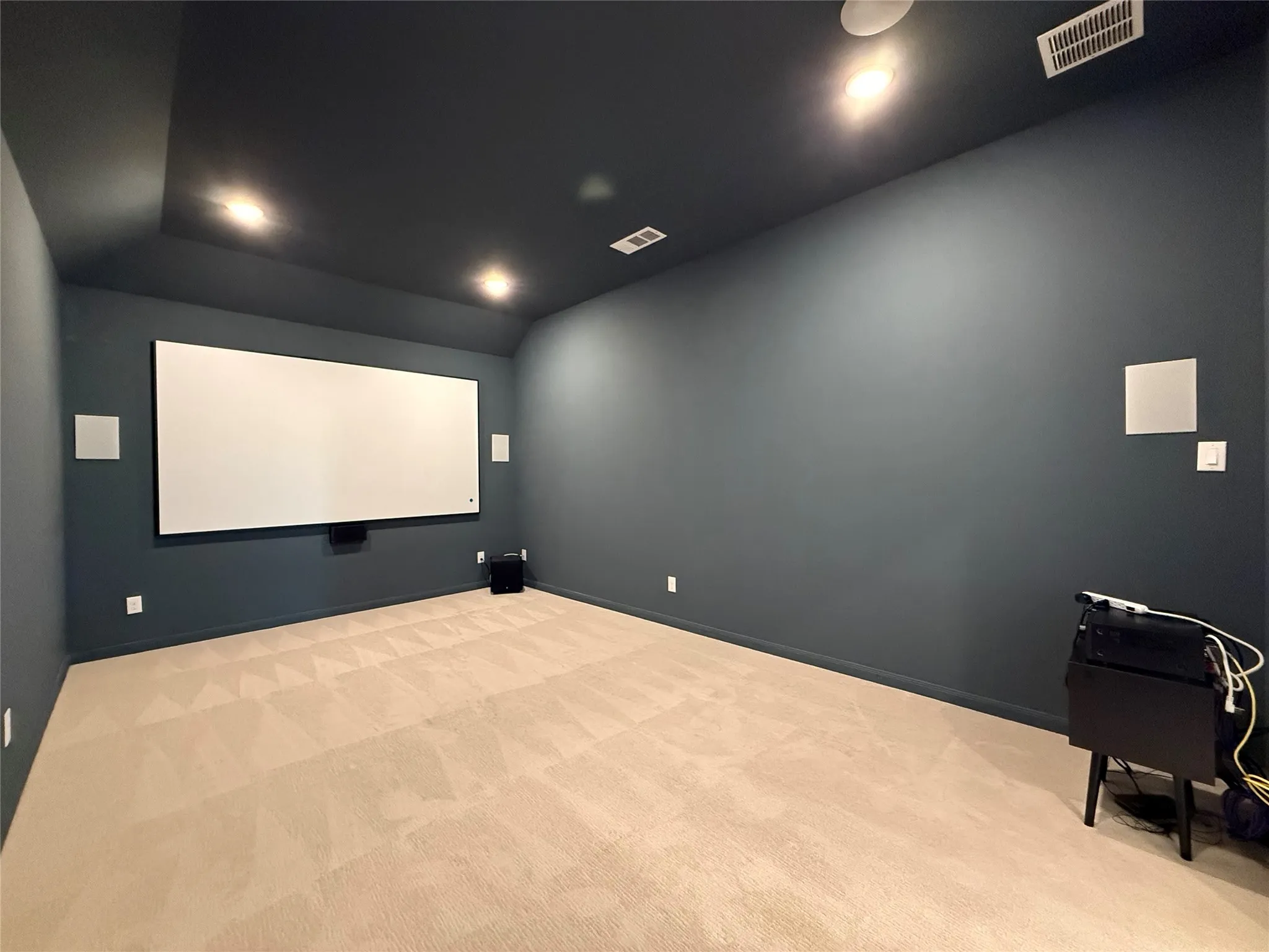 Interior room featuring a tray ceiling, recessed lighting, wall-mounted projection screen, built-in wall speakers, and neutral carpeting