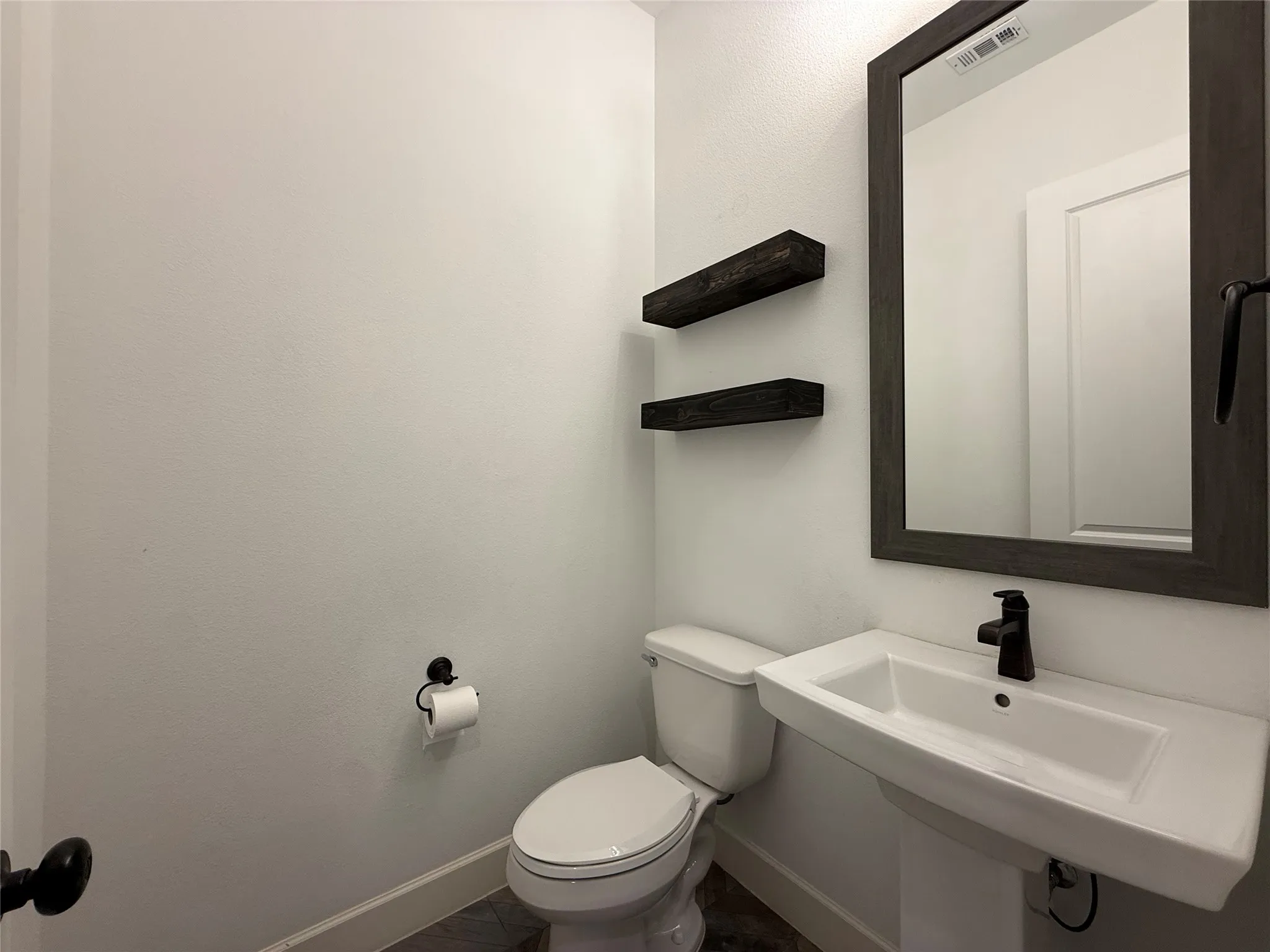 Pedestal sink with matte black faucet, wall-mounted dark wood-finish shelving, and a framed vanity mirror