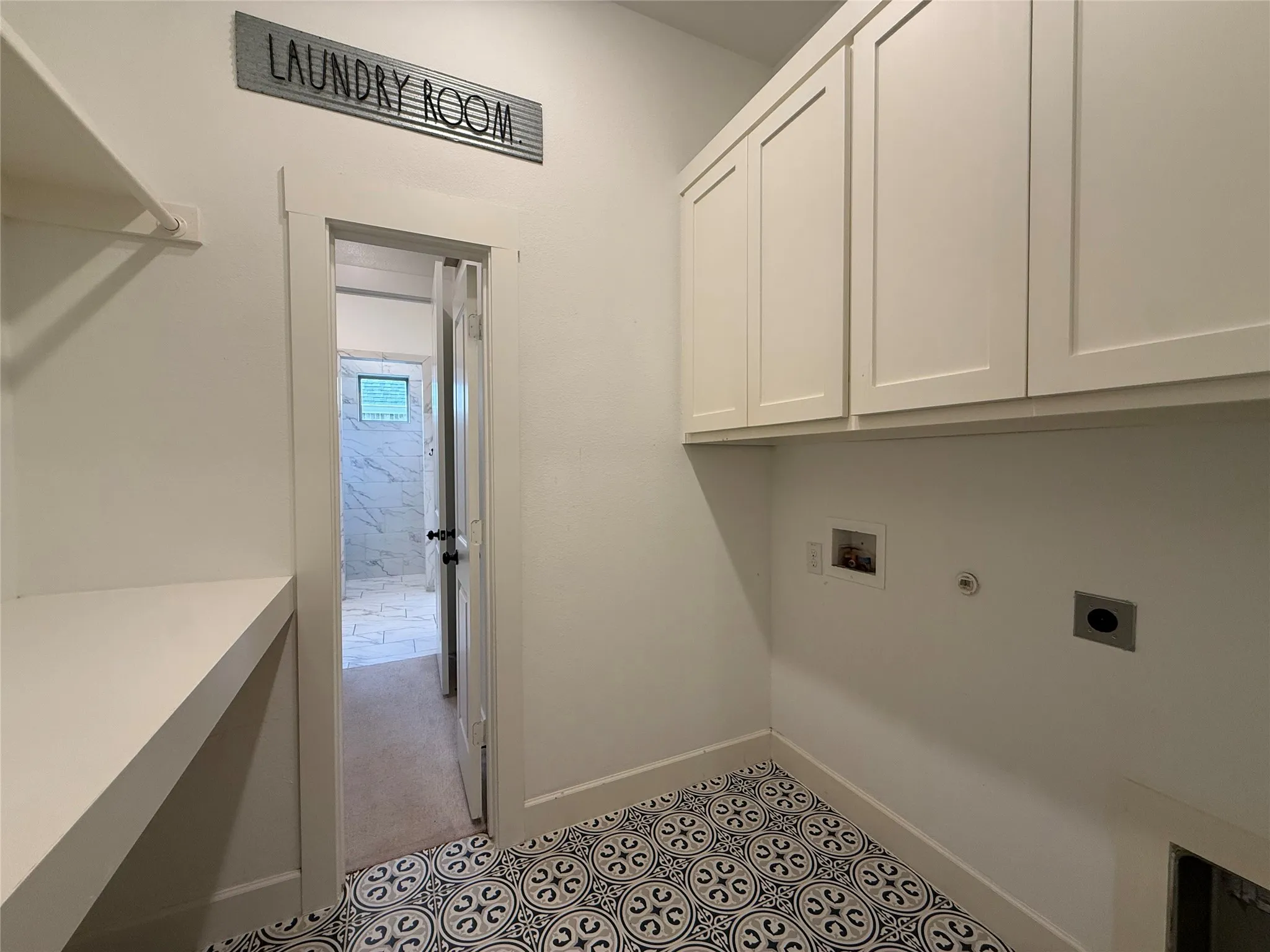 Utility area featuring patterned tile flooring, built-in white upper cabinetry, a fixed shelving unit, and plumbing hookups