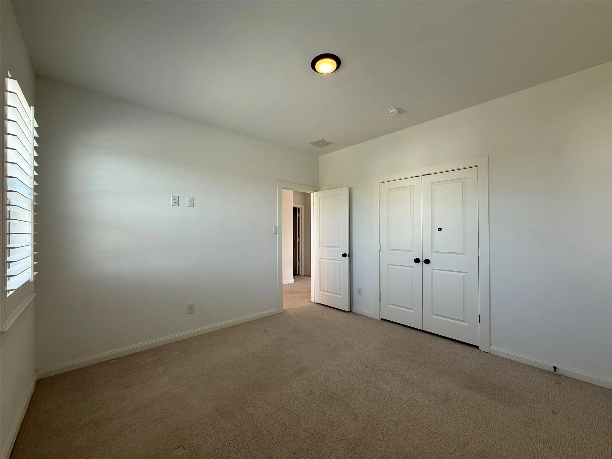 Interior room featuring neutral wall-to-wall carpeting, white walls, plantation shutters, dual-panel closet doors with matte black hardware, and an overhead flush-mount light fixture