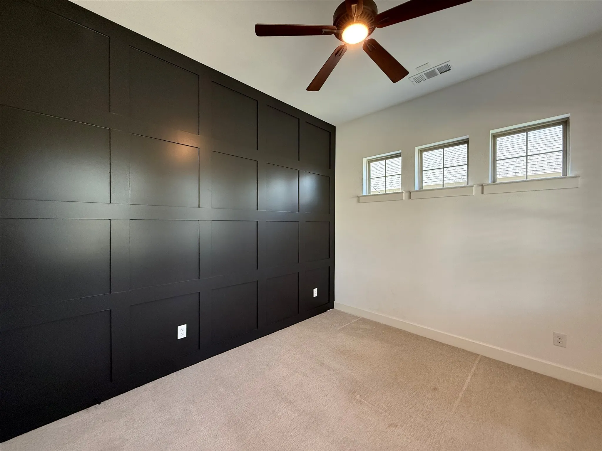 Accent wall featuring black grid molding, neutral carpeting, three high-set windows with grid muntins, and an attached ceiling fan with integrated lighting