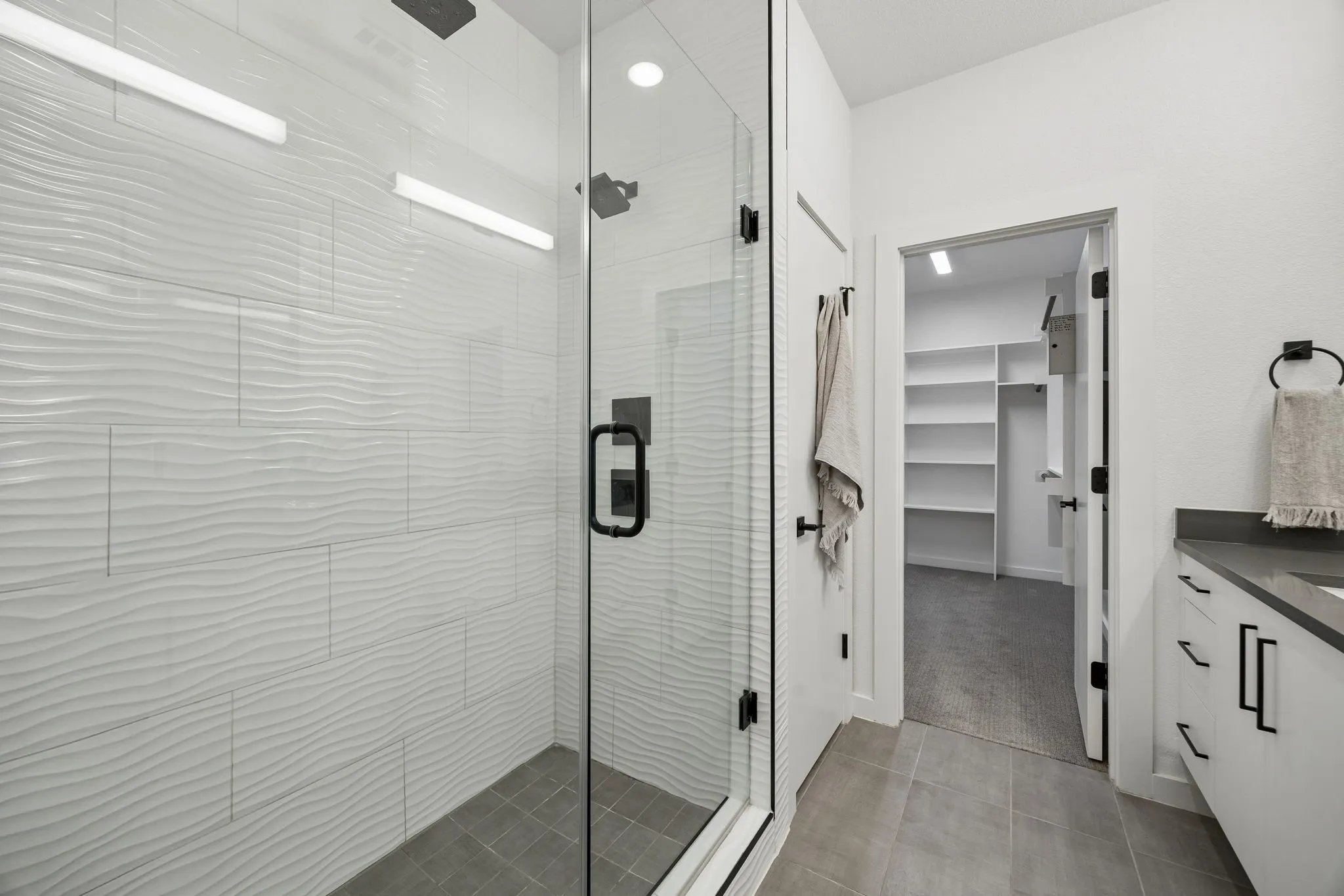 Walk-in shower featuring textured white tile surround, glass enclosure, matte black fixtures, and grey floor tiling, complemented by a white vanity with dark countertop and built-in shelving