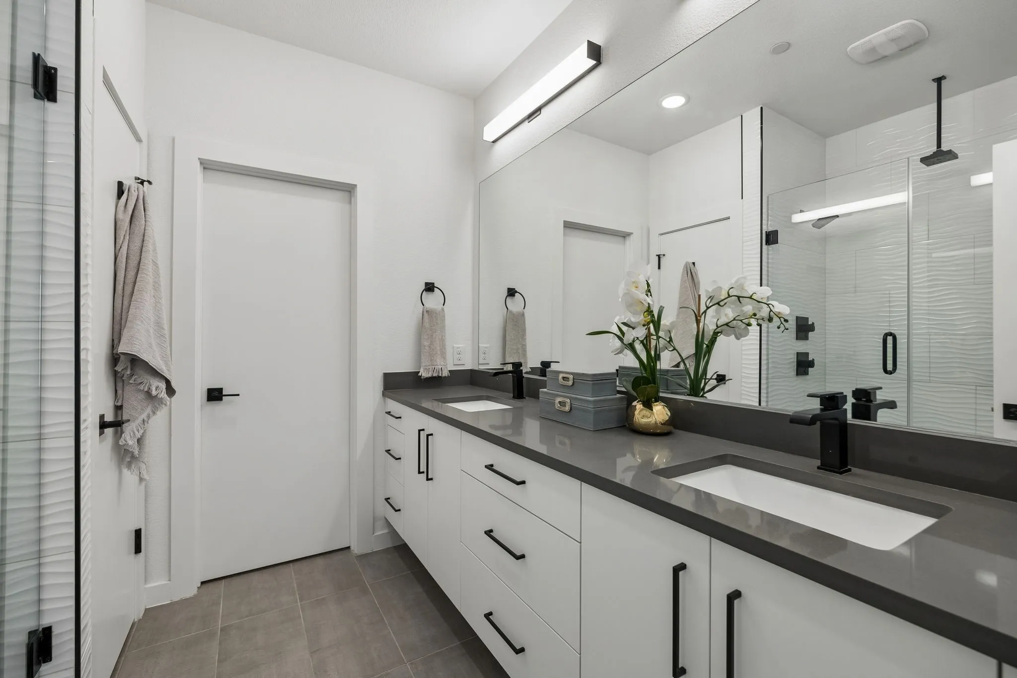 Dual-sink vanity with matte dark hardware, charcoal-toned solid surface countertop, expansive wall-to-wall mirror, tiled flooring, and walk-in shower with glass enclosure