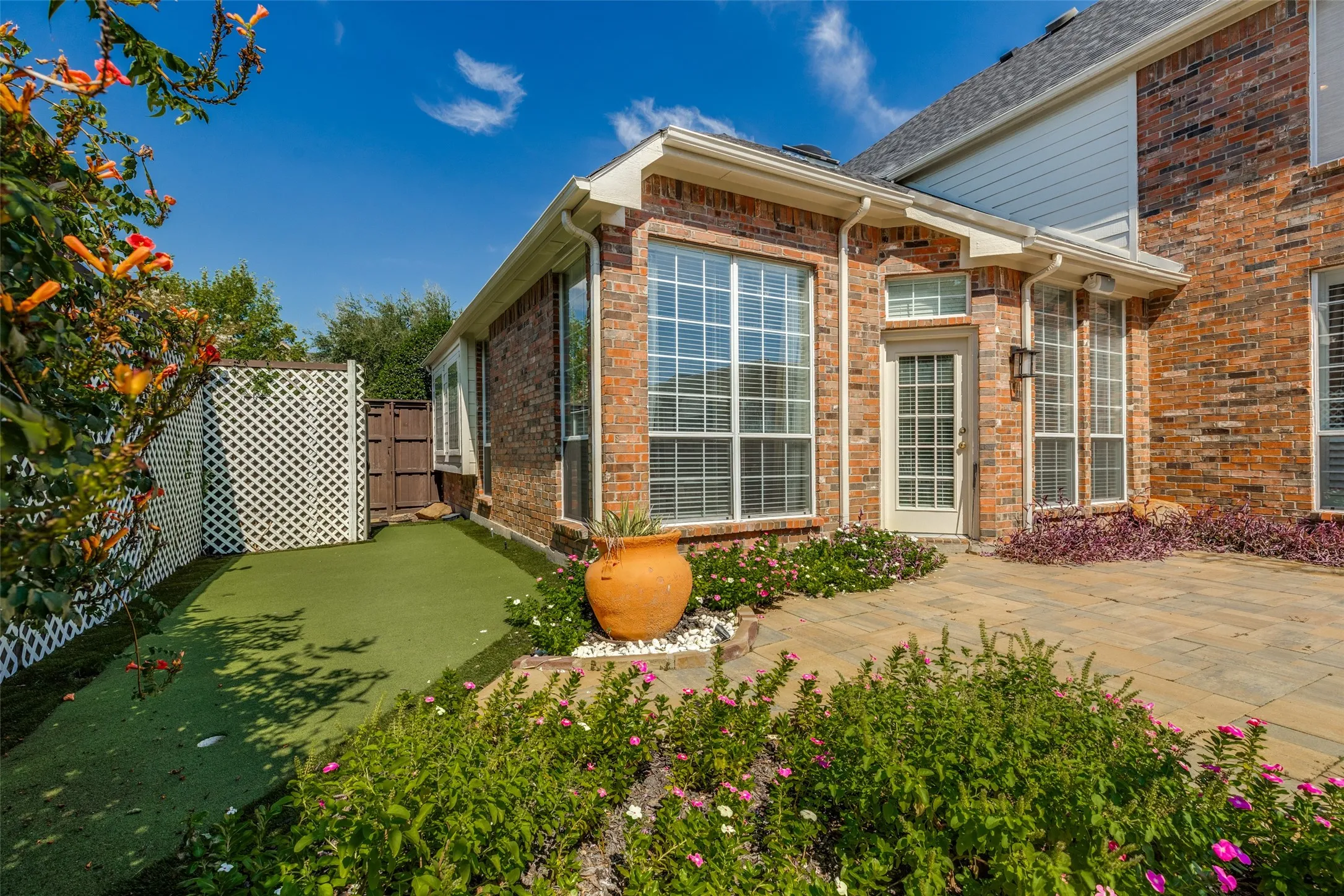 Brick and siding exterior featuring a natural stone patio, synthetic lawn area, lattice privacy screening, and multiple pane-glass windows