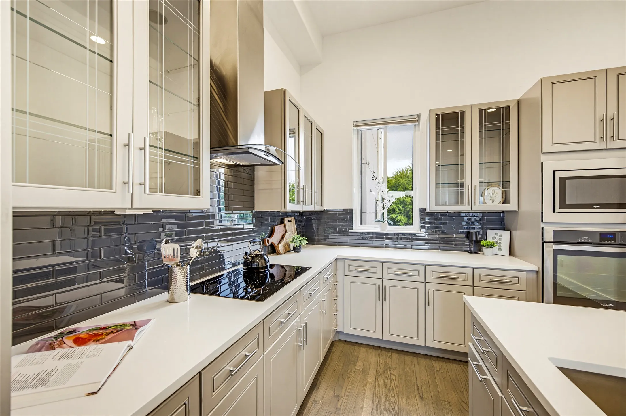 Kitchen featuring light-toned cabinetry with glass-front uppers, solid-surface countertops, dark subway-style tile backsplash, stainless steel range hood, and wood-finish flooring