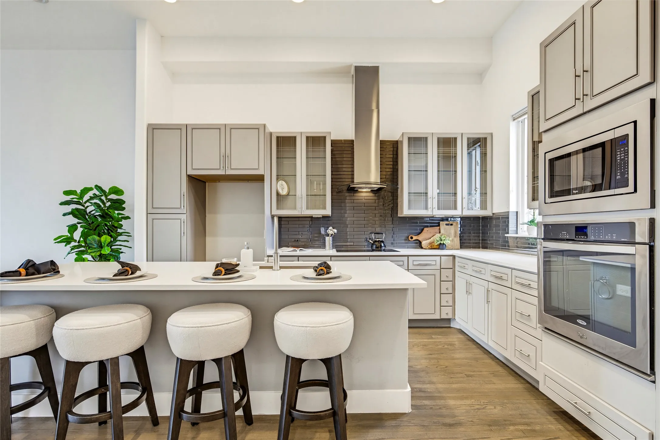 Kitchen featuring light gray shaker-style cabinetry, quartz countertops, a central island with bar seating, stainless steel appliances, and a dark tile backsplash