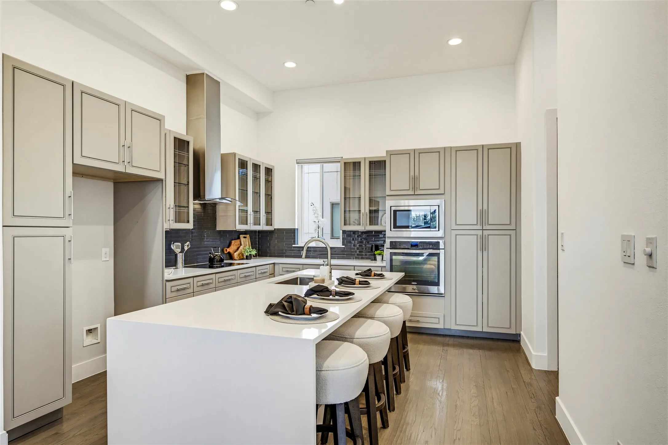 Kitchen featuring light-toned cabinetry with glass-front accents, an expansive quartz-topped island, stainless steel built-in double ovens, a chimney-style range hood, and wood-finish flooring