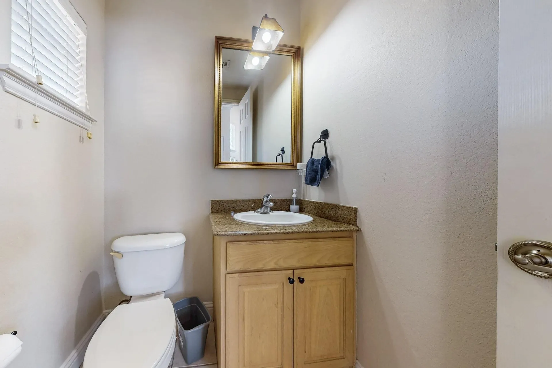 Vanity featuring light wood-finish cabinetry, a stone-composite countertop with an integrated drop-in sink, a wall-mounted light fixture, and a framed mirror