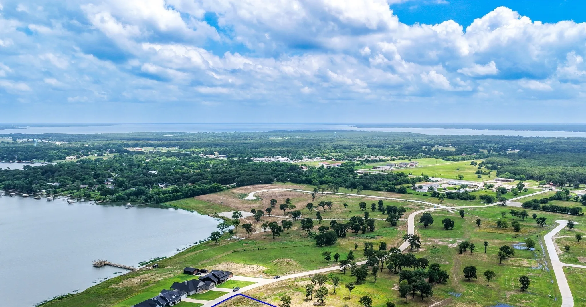Aerial perspective of waterfront parcel featuring expansive grassy terrain, mature trees, a private wooden dock, and paved road access