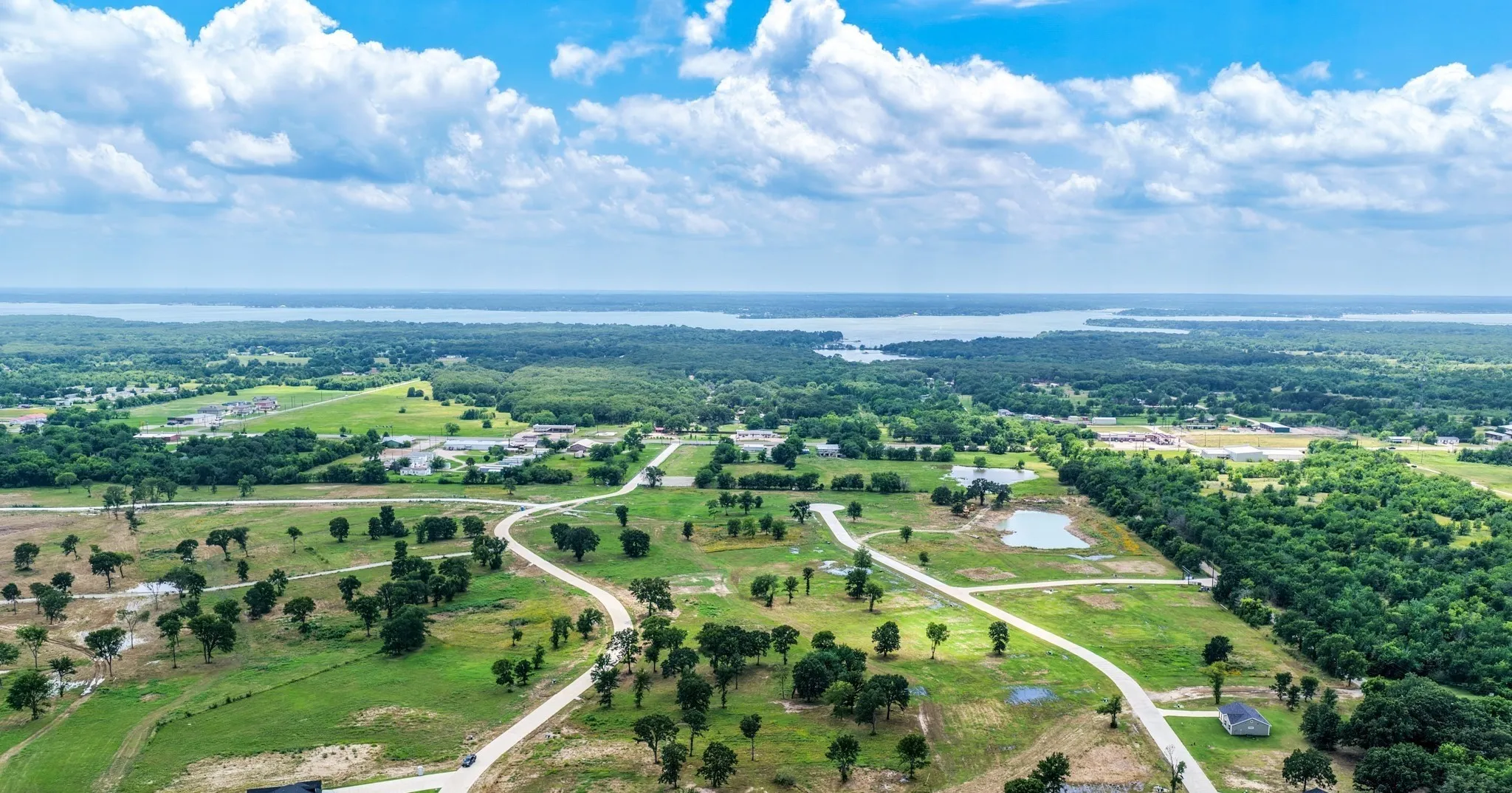 Expansive acreage featuring cleared pasture land, mature trees, paved roadways, and a water backdrop