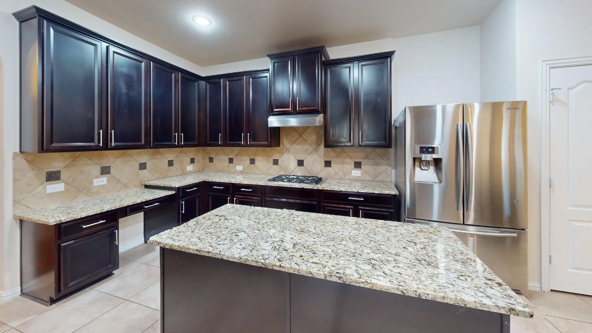 Kitchen featuring dark-finish cabinetry, natural stone countertops, a central island, tile backsplash with decorative accents, and stainless steel appliances
