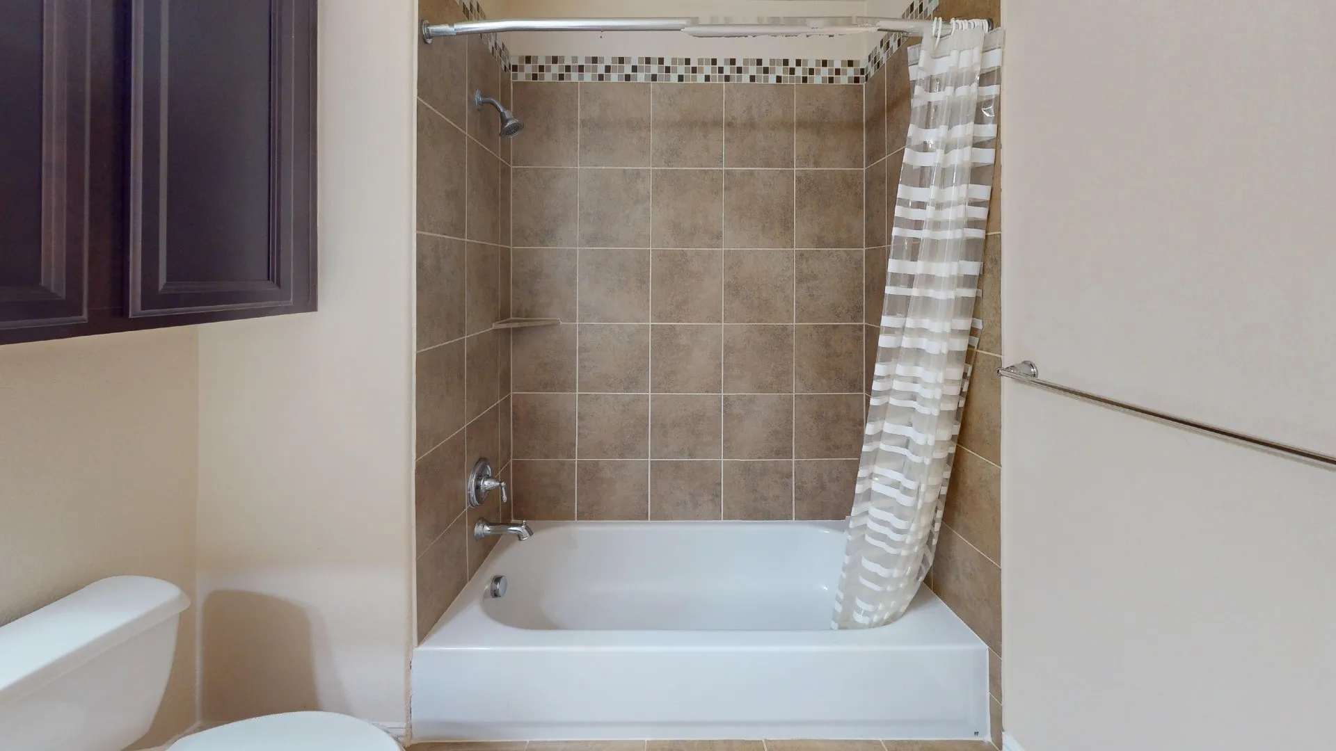 Bathtub and shower combination featuring earth-toned wall tile, a decorative mosaic accent border, integrated soap ledge, and dark wood-finish cabinetry