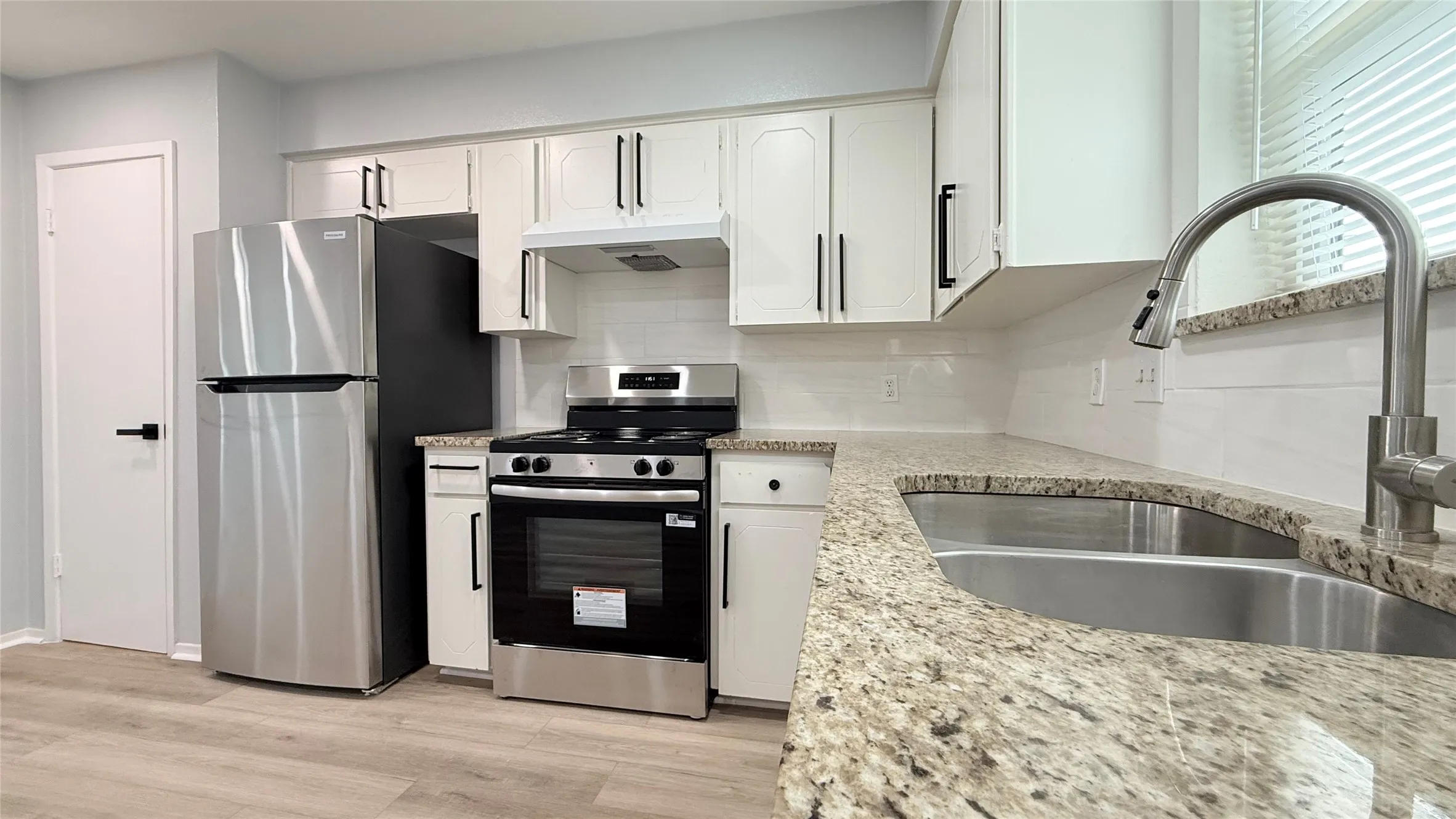 Kitchen featuring white shaker-style cabinetry, stone-patterned countertops, stainless steel appliances, wood-finish flooring, and a dual-basin stainless steel sink with a high-arch pull-down faucet