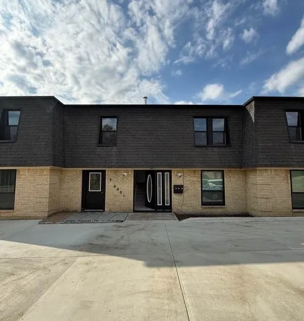 Exterior facade featuring tan brick masonry, dark shingle siding, rectangular windows, and a paved concrete parking area
