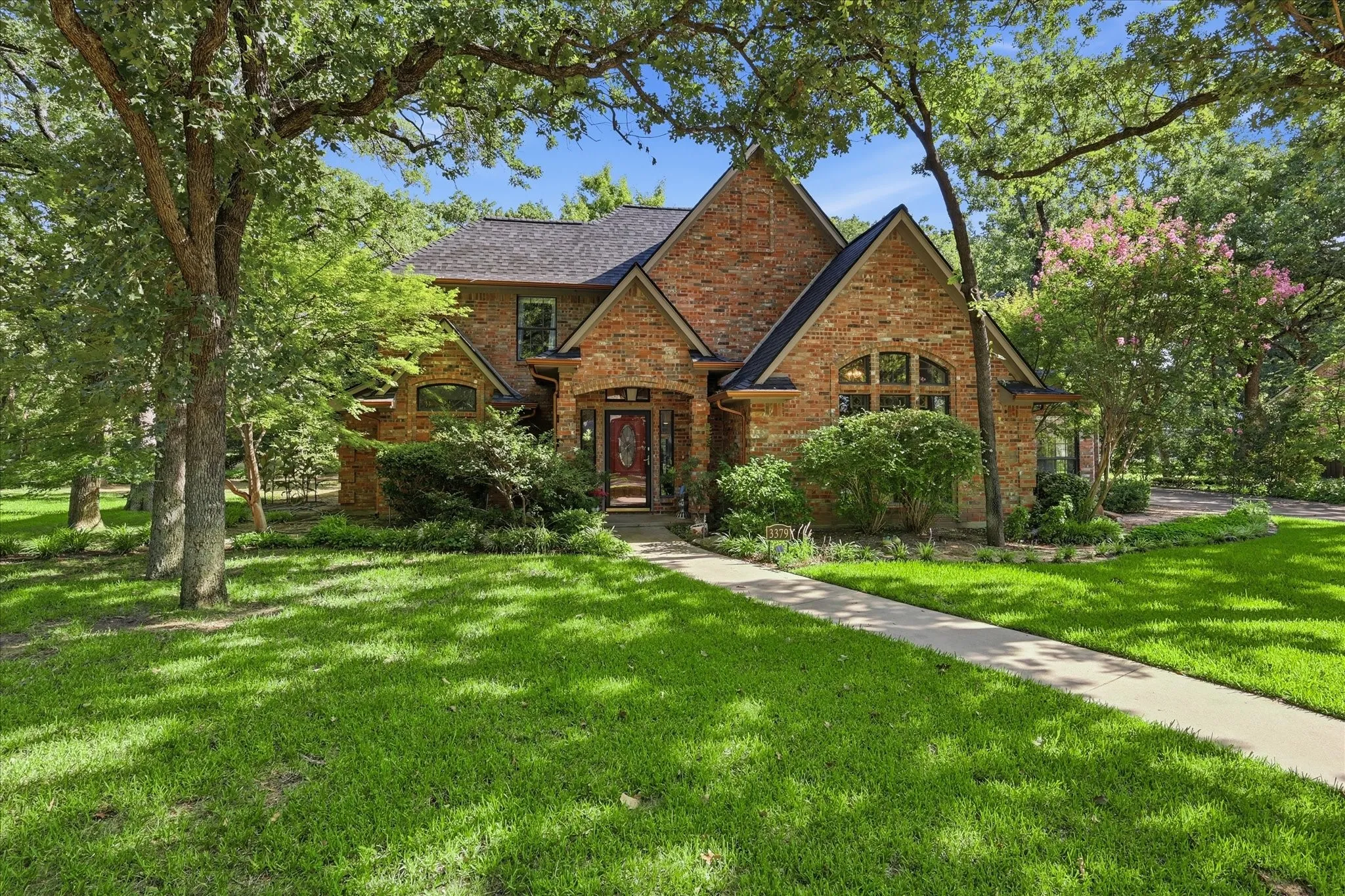 Traditional brick exterior featuring a steeply pitched roof, arched windows, covered entrance, and mature trees on a landscaped lot
