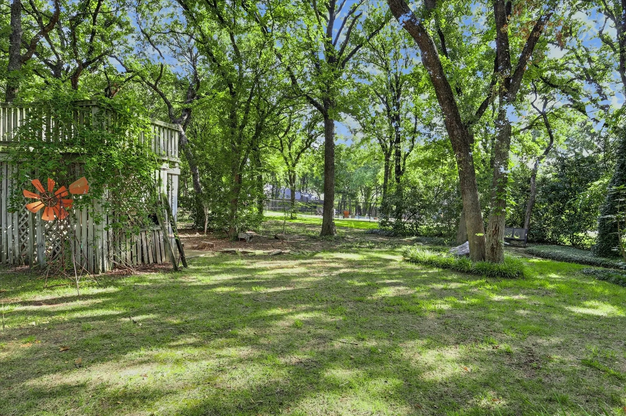 Expansive backyard featuring mature shade trees, a grass lawn, decorative metal windmill, and tiered wooden fencing