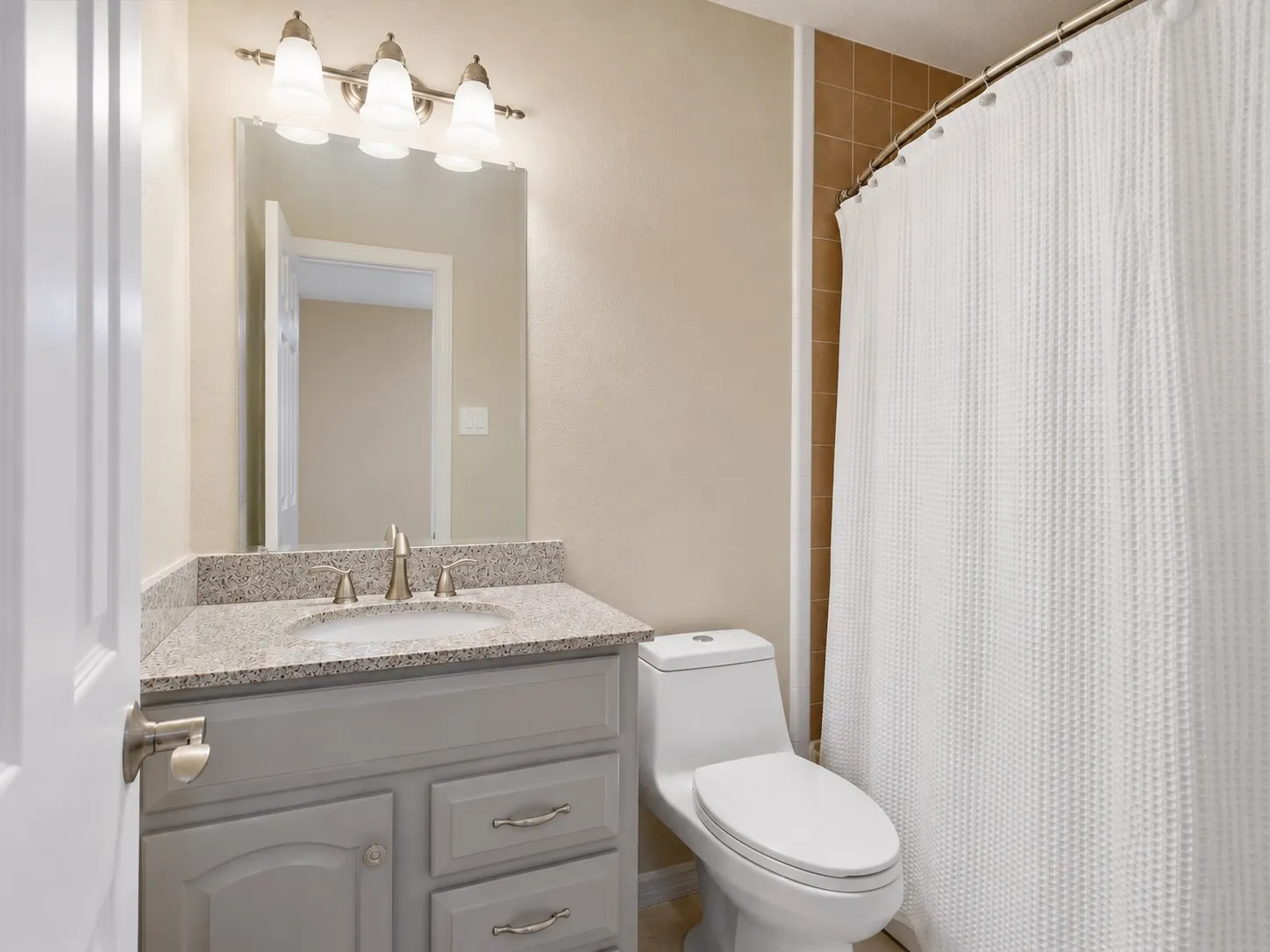 Bathroom featuring a grey vanity with stone-finish countertop, undermount sink, brushed nickel hardware, three-light fixture, and a curved shower rod
