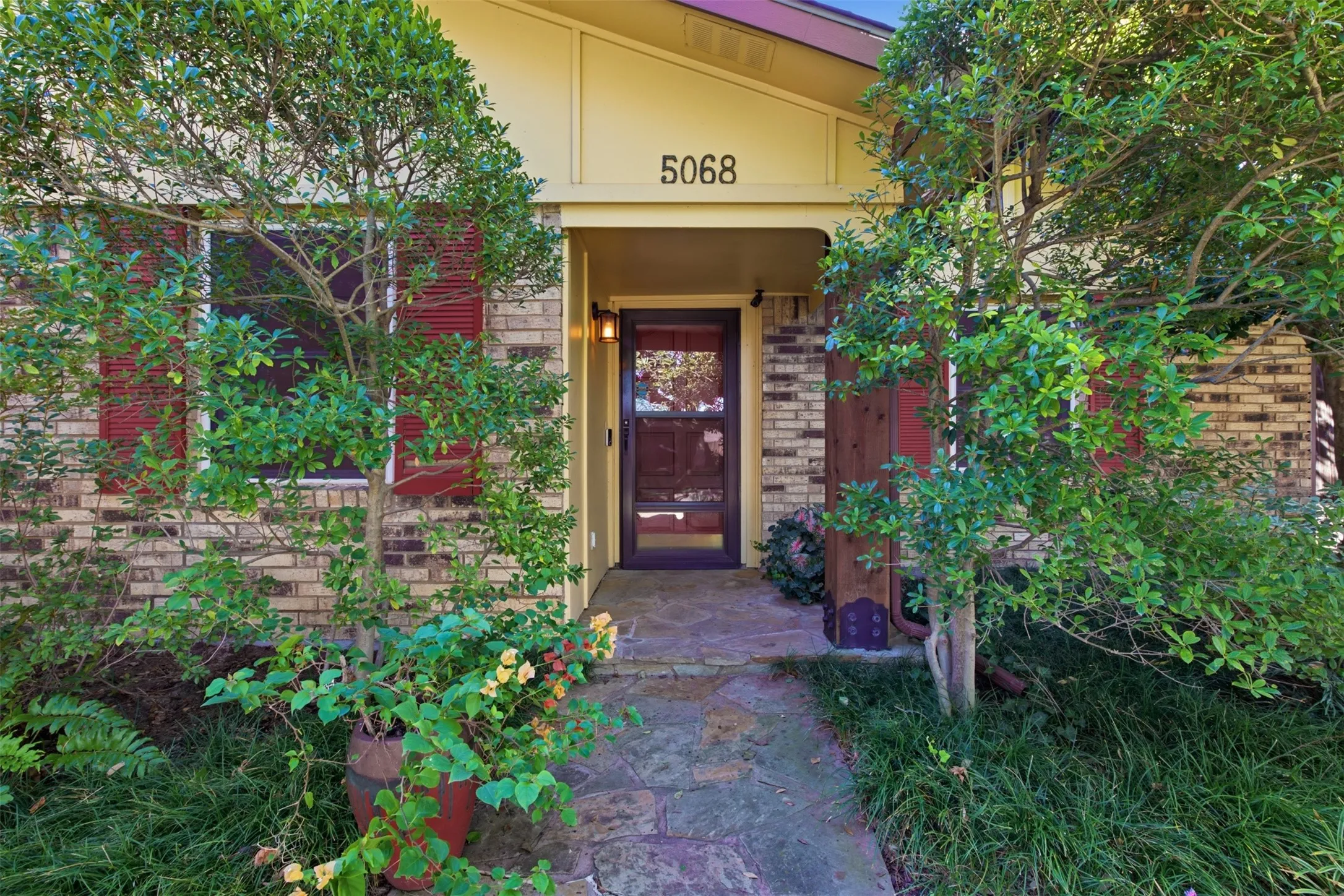 Brick exterior with red shutters, stone pathway, covered entry, and lush landscaping