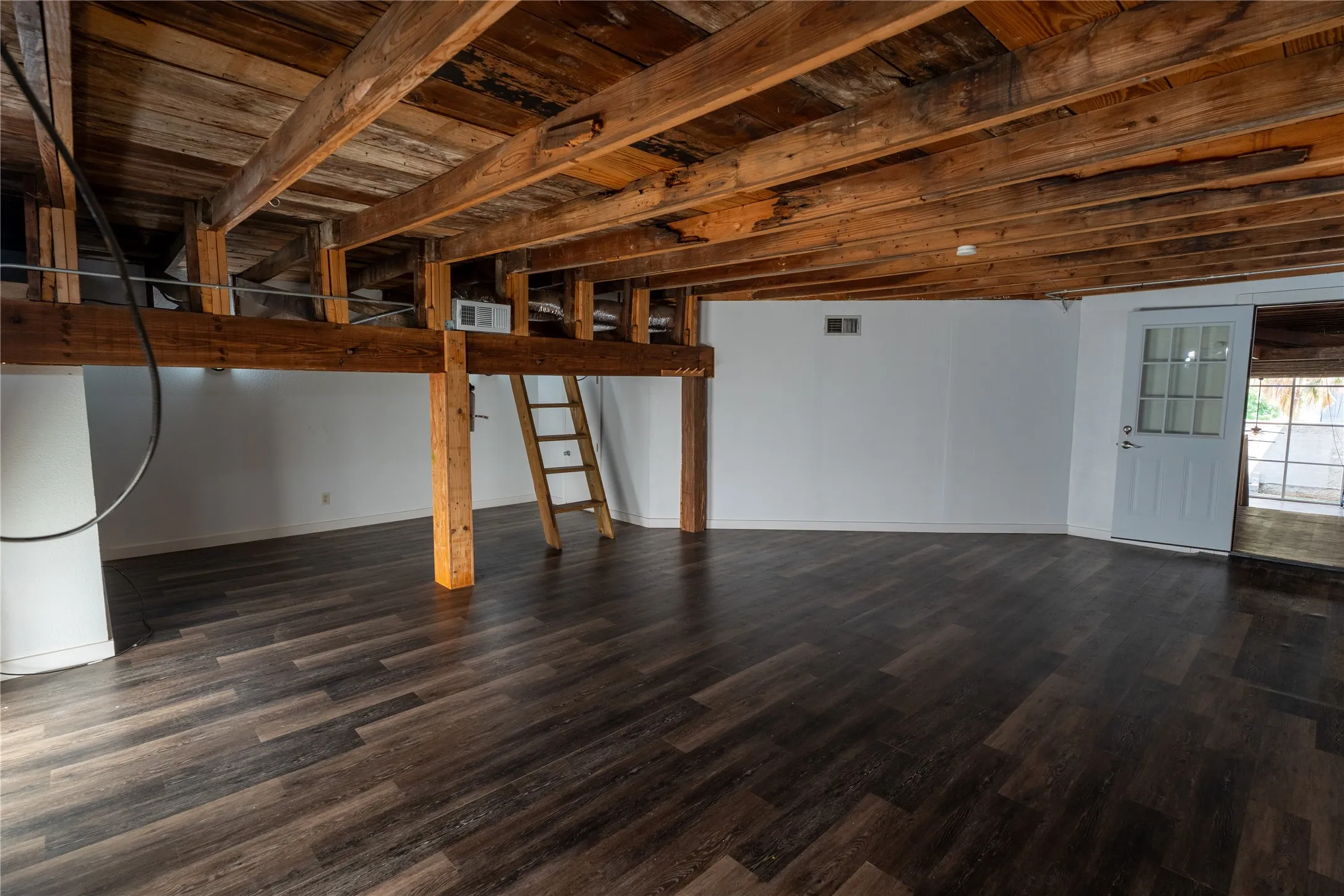Interior space featuring exposed wooden ceiling beams and support posts, wood-finish flooring, white walls, and a multi-pane glass-panel door
