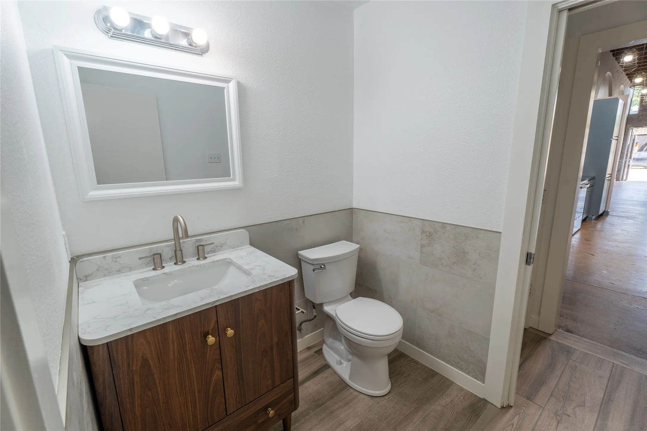 Vanity featuring a stone-patterned countertop, under-mount sink, and wood-finish cabinetry, complemented by wall-mounted lighting, a rectangular mirror, and neutral tile wainscoting